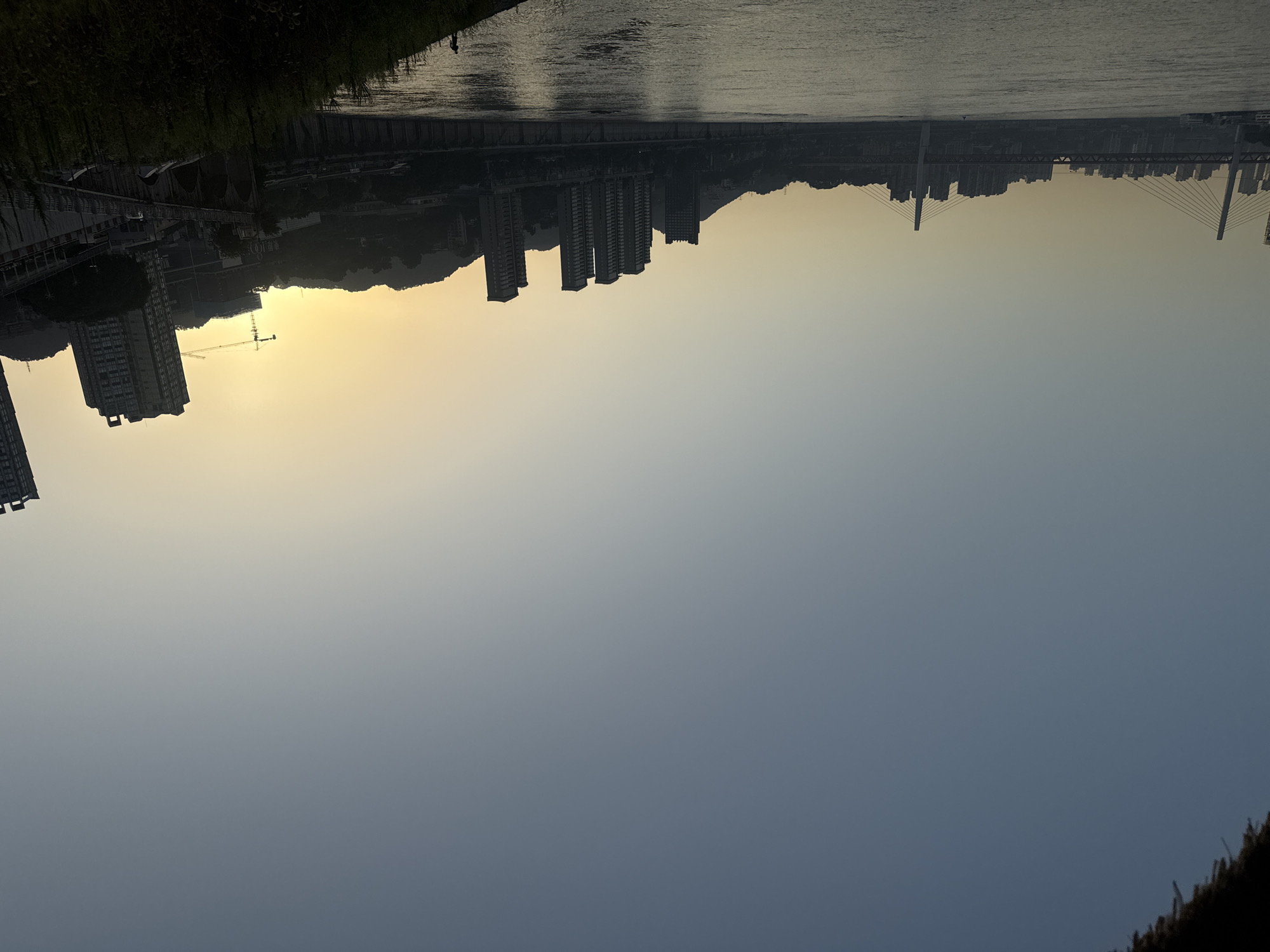 This image depicts a panoramic view of the Yangtze River flowing through the city of Chongqing, China, as seen from the Nan'an District. The scene is captured during either sunrise or sunset, with a warm, soft glow illuminating the sky and casting long shadows over the cityscape.
In the foreground, the wide expanse of the Yangtze River occupies the lower half of the frame, its surface rippling gently. A lone figure, silhouetted against the fading light, can be seen standing on the grassy embankment on the right side of the river.
Across the river, the Nan'an District skyline rises, characterized by a dense collection of modern high-rise buildings. The architecture of these buildings is varied, with some featuring sleek, glass facades and others appearing more traditional. A prominent cable-stayed bridge spans the river, connecting different parts of the city. Its two tall pylons rise majestically, supporting the intricate network of cables that hold up the bridge deck. Further into the distance, more buildings and structures are visible, nestled against the backdrop of rolling hills.
A construction crane can be observed on the right side of the frame, suggesting ongoing development within the city. The overall atmosphere is serene and tranquil, with the natural beauty of the river and the man-made structures of the city harmoniously coexisting. The sky is a gradient of soft blues and yellows, transitioning from a clear, pale blue at the top to a warm, golden hue near the horizon.