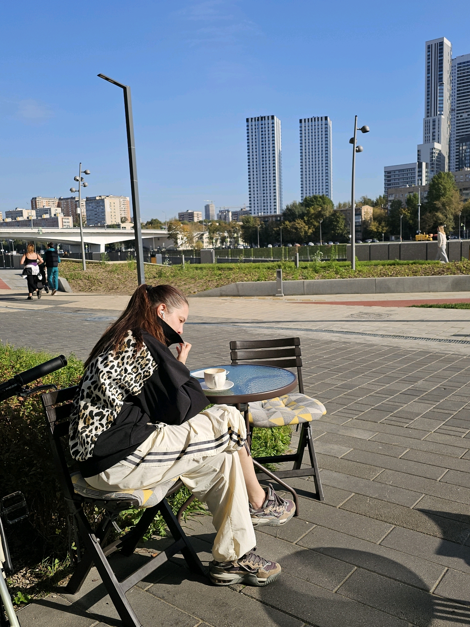 In Moscow, Russia, a woman is seated at an outdoor cafe table. She is wearing a black and leopard-print jacket, cream-colored sweatpants with black stripes, and athletic shoes. She has earbuds in and is holding her phone to her ear, appearing to be on a call. A cup of coffee sits on the table in front of her. In the background, several modern high-rise buildings are visible, along with trees and a clear blue sky. The cafe is located on a paved area with other patrons walking in the distance.
