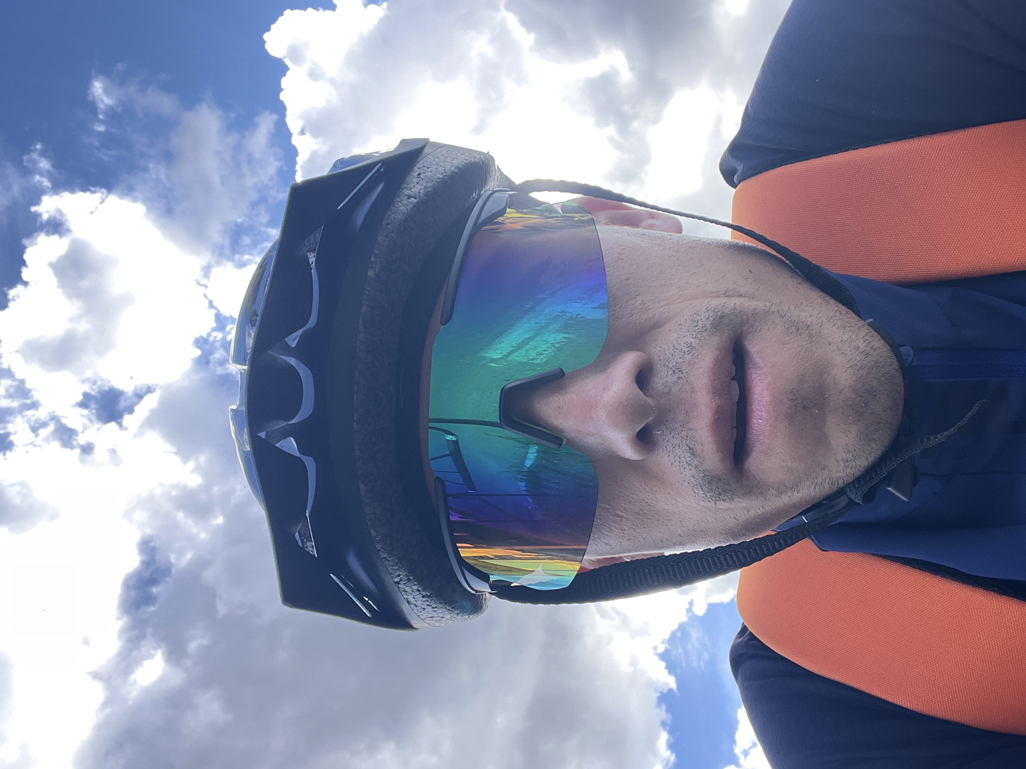 A man is taking a selfie outdoors. He is wearing a black cycling helmet and iridescent sunglasses that reflect a landscape with trees and a body of water. He also wears a dark blue shirt and an orange garment. The background is a blue sky with white clouds. The location and specific city in Russia cannot be determined from this image alone.
