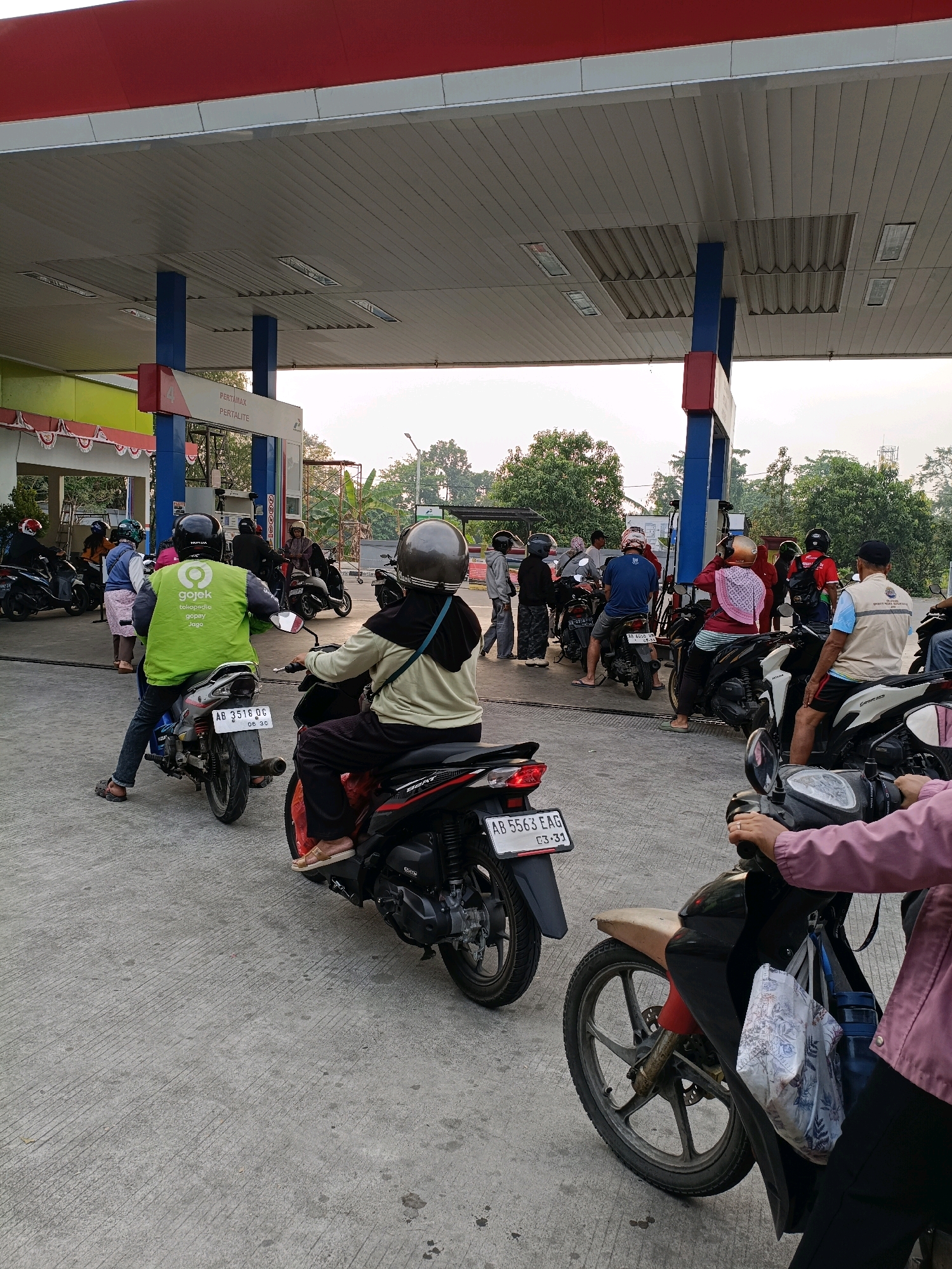 The image depicts a queue of motorcyclists at a Pertamina gas station in Maguwoharjo, Indonesia. The gas station has a red and white canopy supported by blue and red pillars. Multiple motorcycles are lined up, with several individuals wearing helmets and casual clothing. One person in the foreground, wearing a green vest with "Gojek" and "Tokopedia" logos, is on a motorcycle. Another person in the middle ground, wearing a light brown jacket and a dark headscarf, is also on a motorcycle. In the background, more people and motorcycles are visible, along with trees and buildings. The ground is paved with concrete. The overall scene suggests a typical day at a busy gas station.
