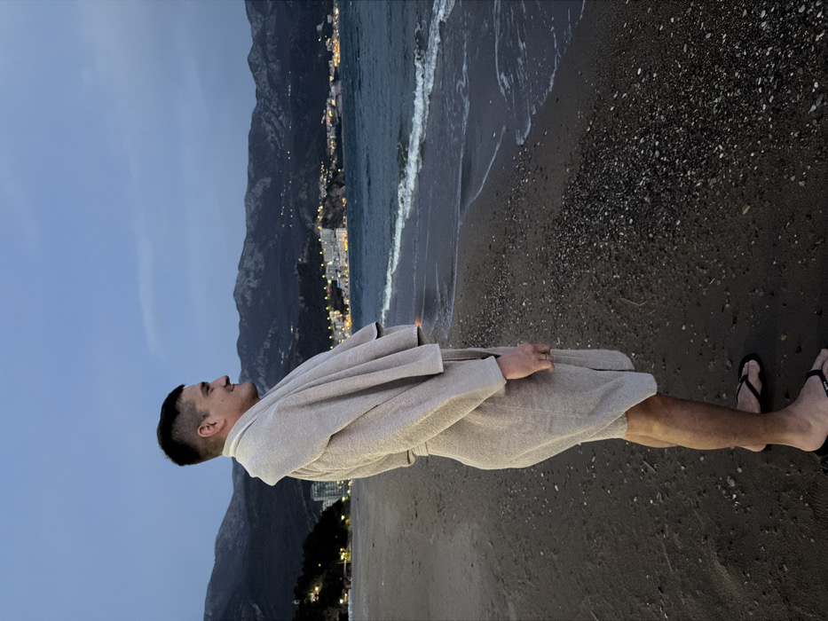 The image displays a male individual standing on a sandy and pebbly beach in Boreti, Montenegro, during twilight conditions. The subject, positioned in profile facing right, is wearing a light beige textured bathrobe, with a matching towel draped over his shoulders. He has short, dark hair and a subtle smile. A ring is present on his left ring finger, and he is wearing black flip-flops. To the subject's right, the dark blue-green sea extends, featuring small waves breaking near the shoreline. In the background, a coastal town is visible with numerous illuminated buildings, casting light reflections on the water. Behind the town, dark, imposing mountains rise against a blue-grey sky, with scattered artificial lights visible on their slopes.