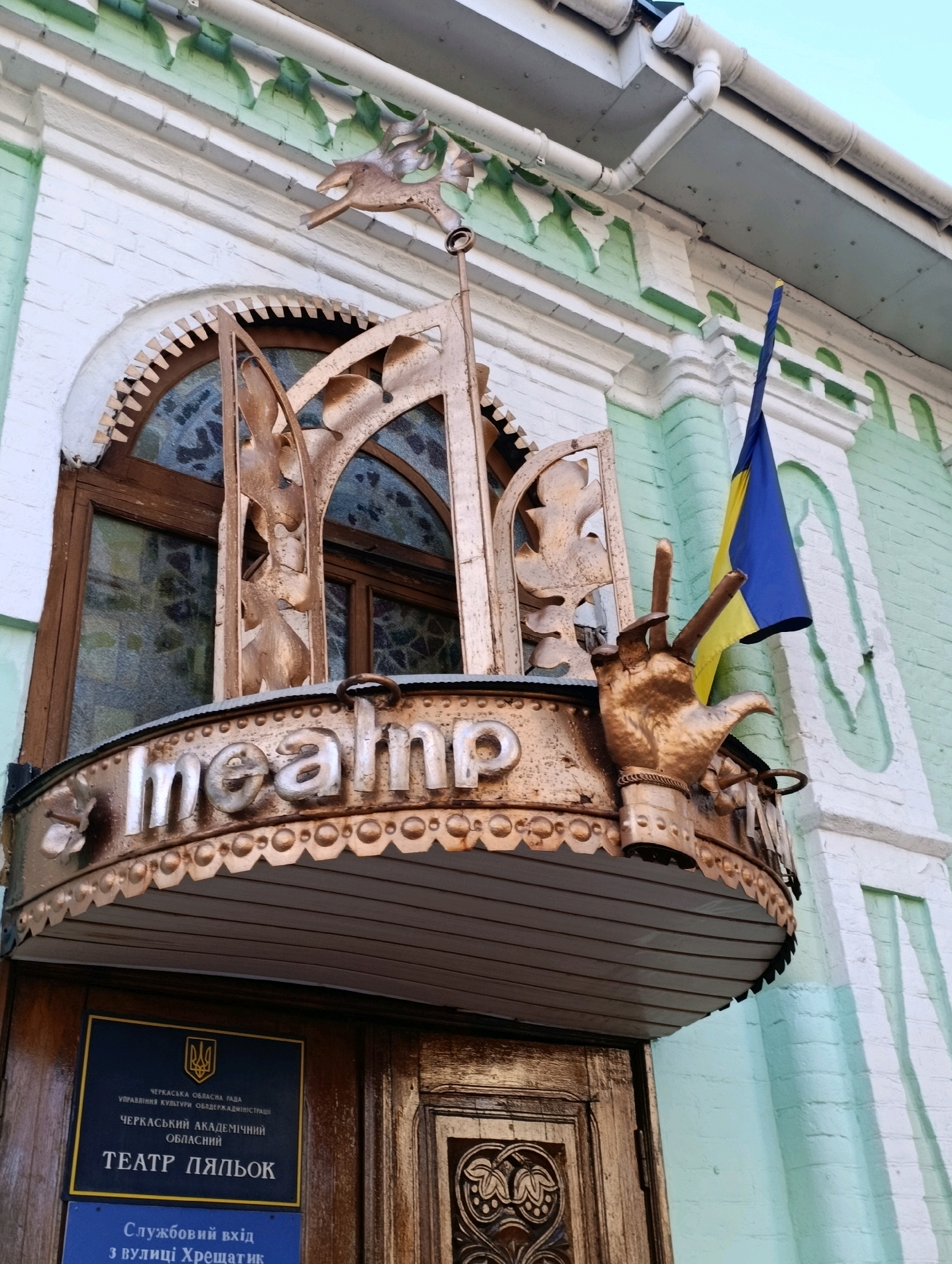 The image shows the exterior of the Cherkasy Academic Puppet Theater located at 4 Khreshchatyk Street in Cherkasy, Ukraine. The building features a light green and white facade with decorative elements above an arched window. A metal balcony with ornate golden detailing, including the words "mealnp" and a large sculpted hand, is visible above the entrance. A Ukrainian flag is mounted on the balcony. Below the balcony, a dark blue sign with text in Ukrainian is affixed to the building, along with a smaller blue sign. The entrance door is made of dark wood with carved detailing. The overall impression is that of a historical building with theatrical elements.
