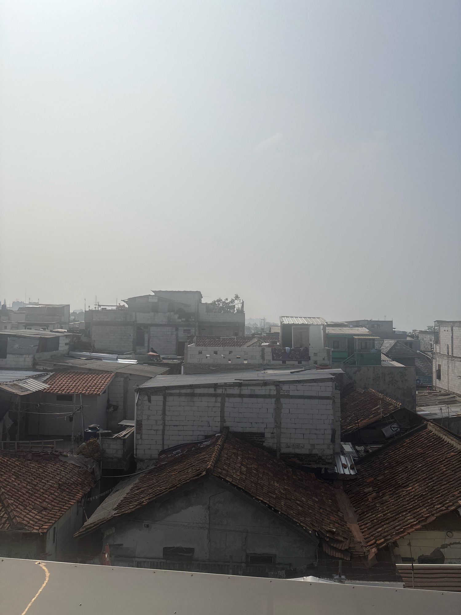 The image shows a densely populated urban area, likely a residential neighborhood, under a hazy sky. The scene is dominated by rooftops of various materials and construction styles, suggesting a mix of older and newer buildings. Many of the structures appear to be made of concrete blocks and corrugated metal, with some featuring traditional terracotta roof tiles. The buildings are tightly packed together, creating a somewhat chaotic but uniform landscape. In the distance, taller buildings and possibly industrial structures are visible, indicating the presence of a larger city. The overall atmosphere is muted due to the haze, which obscures finer details and softens the light. There are no prominent landmarks or specific identifying features that would pinpoint the exact location within Bandung City, Indonesia, beyond the general urban context. The scene appears to be viewed from an elevated position, looking down on the rooftops.