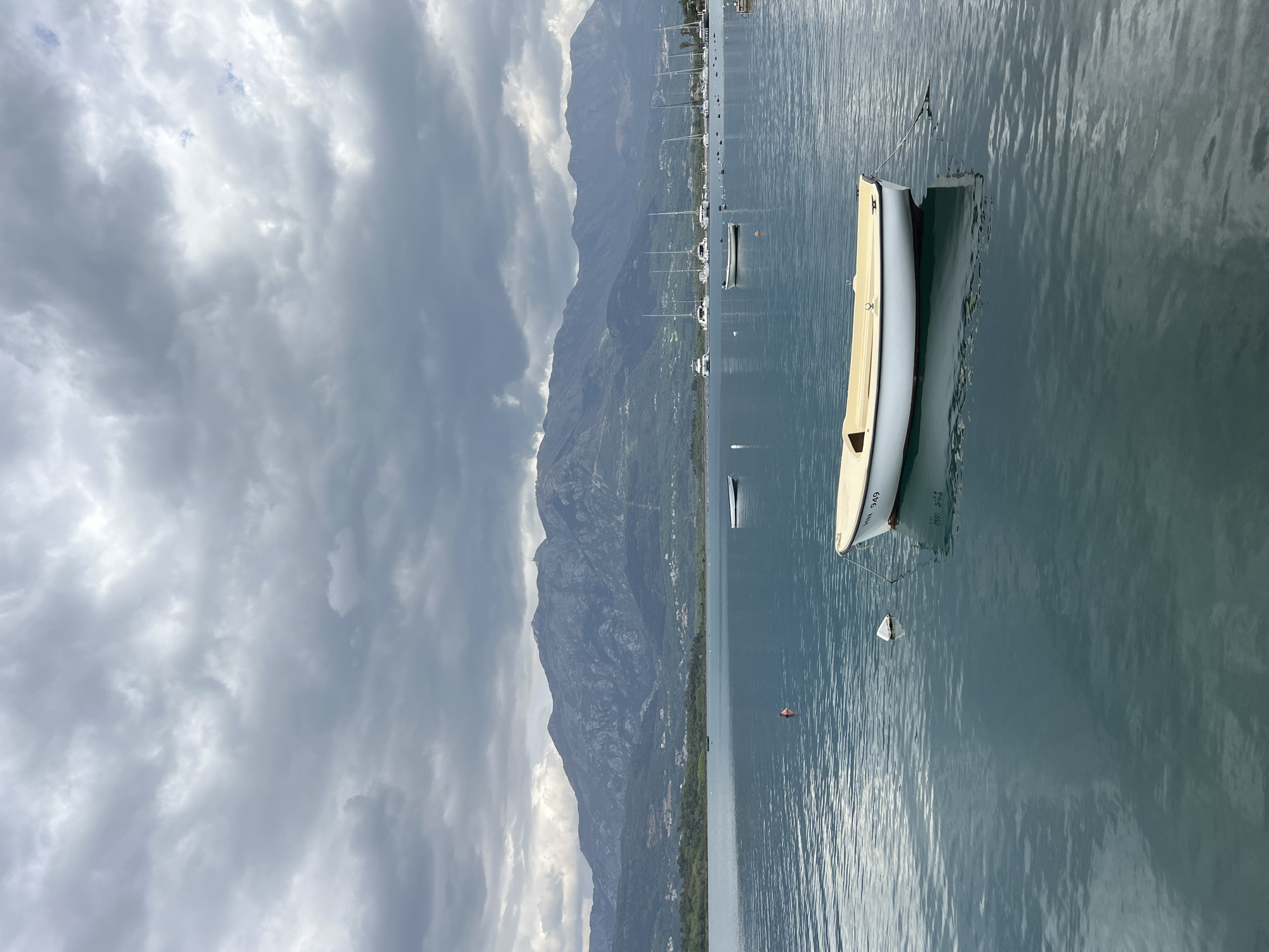 This image captures a serene, overcast day in Đuraševići, Montenegro, showcasing a tranquil body of water against a mountainous backdrop. The composition is horizontally oriented, with a cloudy sky occupying the upper portion and calm water in the lower half.
In the immediate foreground, on the right side of the frame, a light-colored boat, possibly a dinghy or small canoe, is partially visible. Its hull is light grey or off-white, and the number "NN 949" is clearly visible on its side. A small white buoy floats next to it, suggesting the boat is moored. The water around this boat shows gentle ripples and a soft reflection.
Further out on the water, a line of numerous smaller boats, mainly sailboats with their masts upright, are moored across the mid-ground. They appear as dark silhouettes against the water and the distant shore. The water surface itself is mostly calm, displaying subtle ripples that catch and reflect the diffused light from the overcast sky.
The background is dominated by a long, undulating mountain range that stretches horizontally across the scene. The mountains are dark and rugged, with visible patches of green vegetation along their slopes. Scattered buildings and small settlements are nestled at the base of the mountains, along the distant shoreline. The sky above is heavily overcast with thick, grey clouds, which cast a soft, diffused light over the entire scene. There are no people visible, and no overt activity is taking place, contributing to the quiet and peaceful ambiance. The overall impression is one of natural beauty under a muted, cloudy sky.