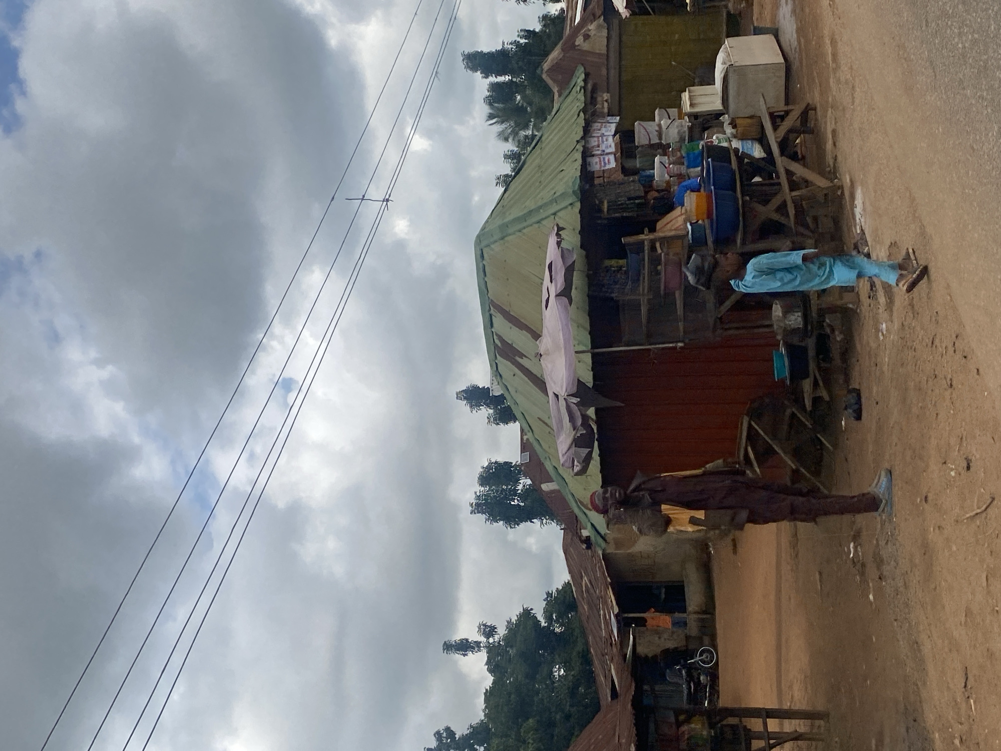 In Tataura, Nigeria, a boy in a blue outfit walks across a dirt road. He carries a dark object on his head, and wears sandals. To his right, an adult male in a brown and yellow outfit stands, facing the boy. Behind them, a market stall with a green roof displays various items, including bowls, buckets, and containers. Further back, a weathered building with a rusted corrugated metal roof stands. Trees with lush green foliage are visible in the background. Power lines traverse the overcast sky.