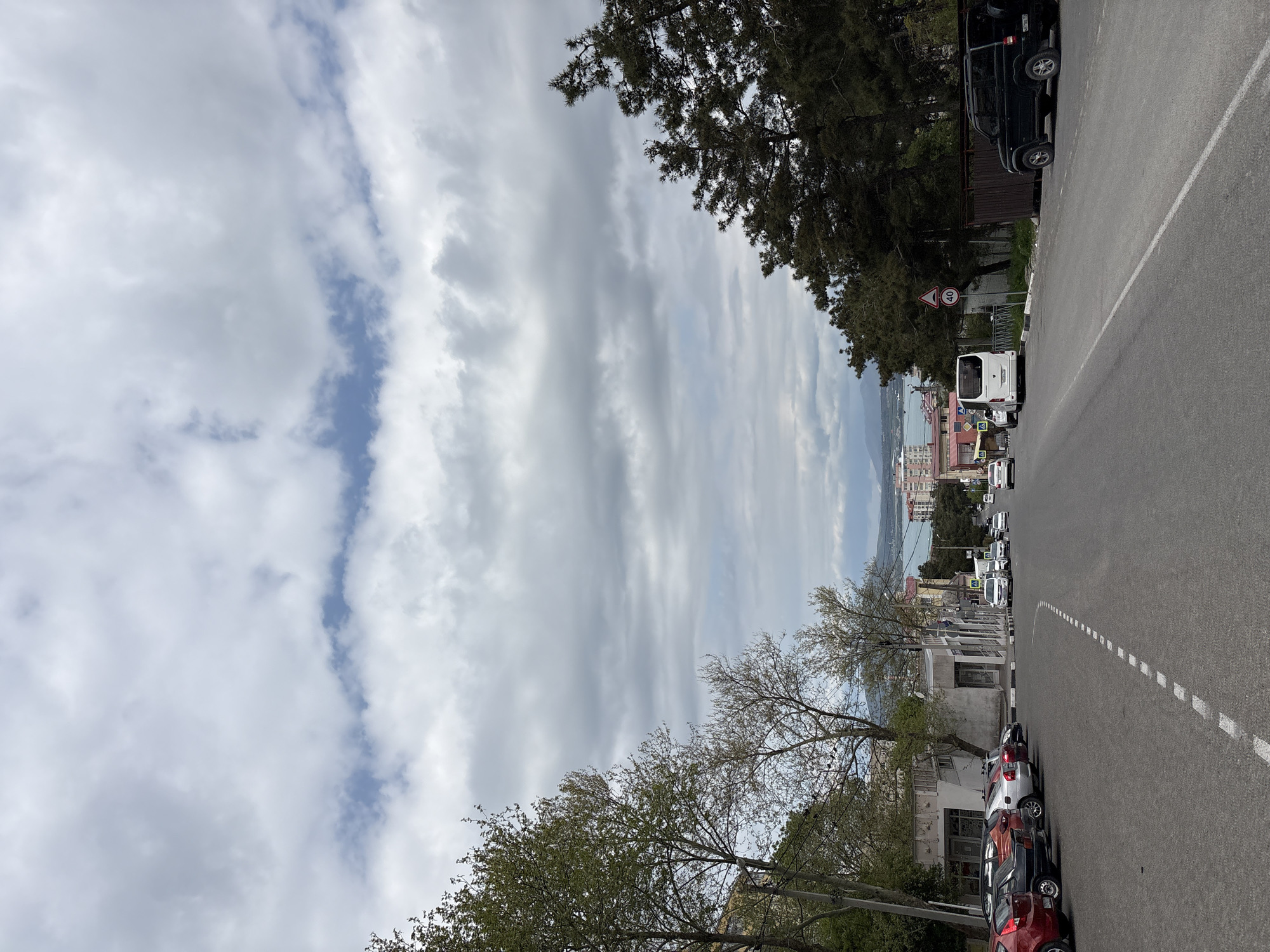 This vertical image depicts a street scene in Gelendzhik, Russia, during what appears to be a cloudy but bright day. The main subject is a paved road that stretches from the foreground into the distance, bordered by various urban and natural elements.
On the right side of the road, a line of cars is parked along the curb, including red, black, and silver vehicles. Behind these cars are buildings, partially obscured by trees that have mostly bare branches. On the left side of the road, there's a pavement and more buildings, with an array of trees, some deciduous and some evergreen, lining the street and extending upwards.
Traffic activity is visible further down the road, with a white bus or large van and other vehicles. A dark SUV is parked under dense evergreen trees on the left. A red circular speed limit sign, displaying "40," is clearly visible on the left side of the road.
In the background, beyond the immediate urban structures and trees, a wide expanse of water, likely the Black Sea, can be seen, with a city and distant hills or mountains on the horizon. The sky is largely covered with elongated, banded clouds, which are light grey and white, but patches of blue sky are visible, especially towards the left. The overall impression is that of a typical street in a coastal town, with no specific event or human activity prominently featured.