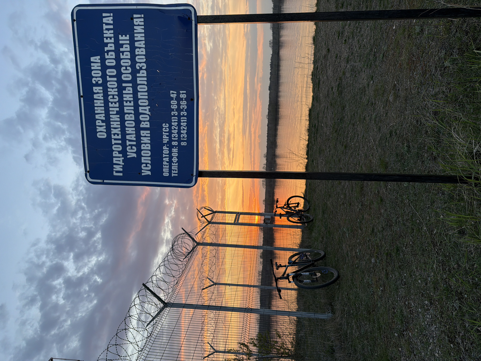 The image depicts a restricted area, likely near a hydroelectric structure, in Chaikovsky, Russia. A blue sign with white lettering warns of restricted access and specifies water use conditions, listing the operator as ChRGS and providing two phone numbers: 8 (34241) 3-60-47 and 8 (34241) 3-36-81. A barbed wire fence runs parallel to the waterfront, with two bicycles leaning against it. The background shows a body of water reflecting a colorful sunset. Tall grass and vegetation are visible on the ground. The sky is a mix of clouds and shades of orange, indicating either sunrise or sunset.