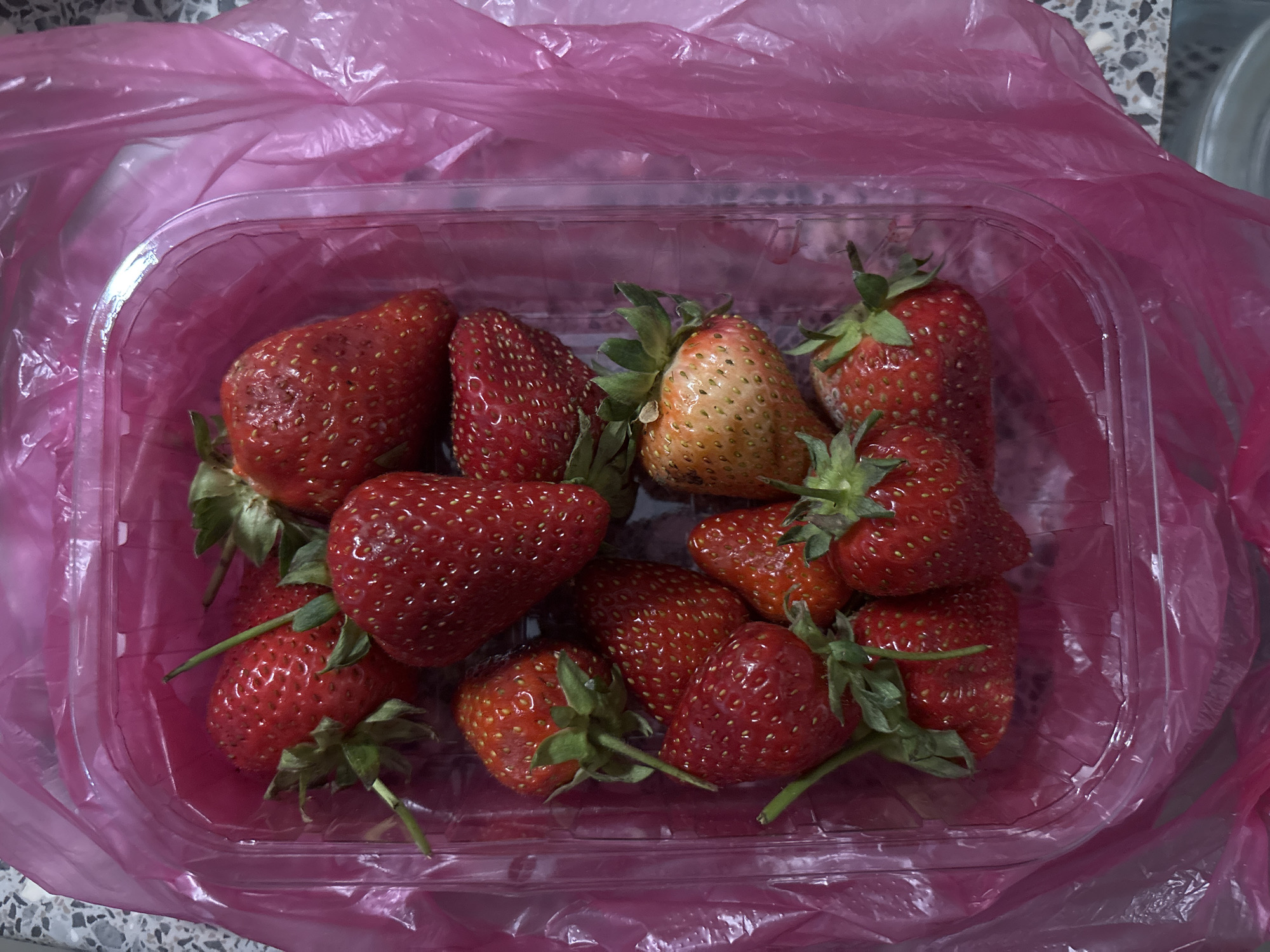 A clear plastic container of strawberries sits inside a pink plastic grocery bag. The container holds approximately a dozen red strawberries with green leafy stems. One strawberry is discolored, showing yellow and brown hues. The surrounding environment appears to be a countertop with a gray and white mottled pattern. Based on the assumption that the photograph adheres to the user's request, the location is Saint Petersburg, Russia.