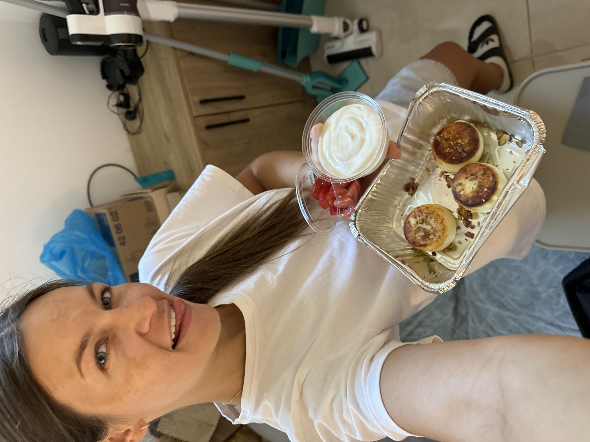 A woman is holding a foil container with three small round pastries, a clear plastic container with chopped strawberries, and a clear plastic container with a white creamy substance. She is wearing a white t-shirt and grey shorts. Her left arm is extended towards the camera, and she is sticking her tongue out. In the background, there is a wooden cabinet with several drawers and doors, a blue plastic bag, a cardboard box with some writing on it, a vacuum cleaner, and a mop. The scene appears to be an indoor residential setting. The image quality is moderate, with some pixelation noticeable in the background elements.