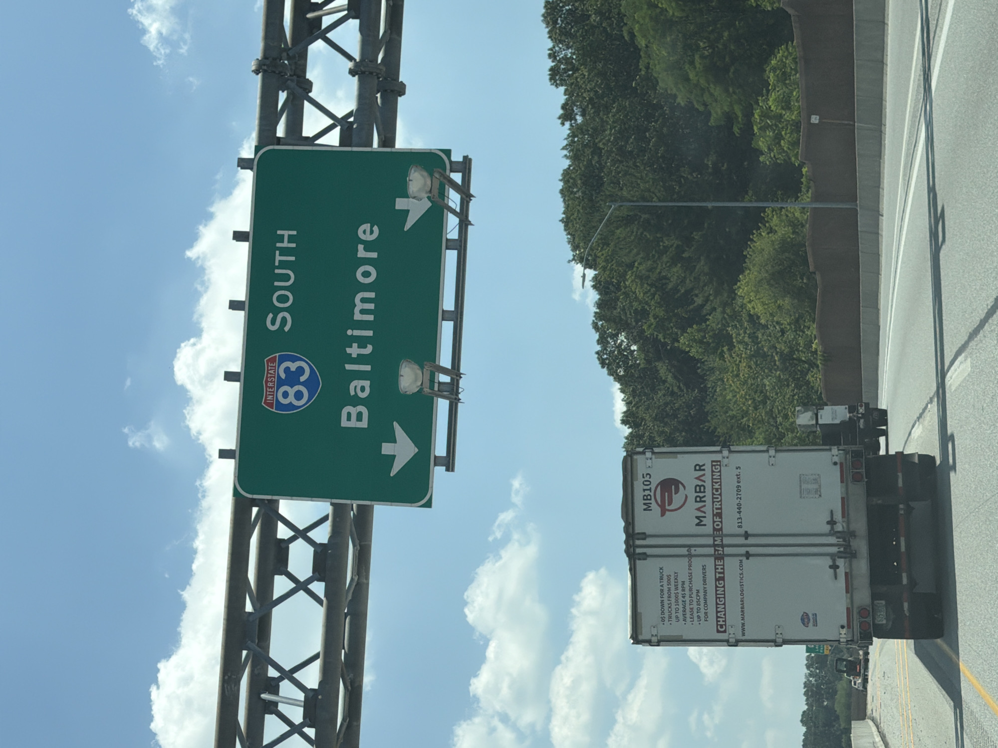 In York Township, United States of America, a green highway sign indicates "INTERSTATE 83 SOUTH BALTIMORE" with downward pointing arrows for both lanes. Below the sign, a white semi-trailer truck with the logo "MARBAR" is traveling on the highway. The truck has text on its side detailing lease and purchase programs for trucks. The scene is under a bright, partly cloudy sky, with lush green trees lining the side of the highway.
