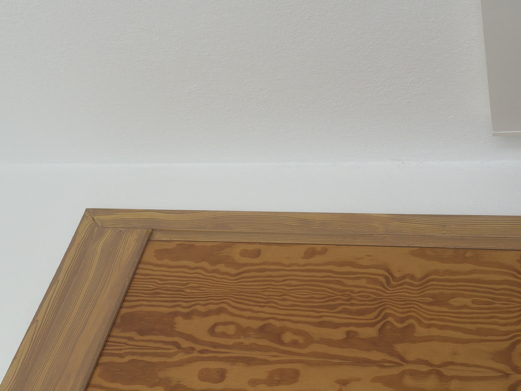 The image shows a close-up view of a wooden surface with a distinct wood grain pattern, framed by a lighter wooden border. The wooden surface appears to be part of a piece of furniture or a wall paneling, with a natural, warm brown tone and visible rings and knots that indicate its natural origin. The wood grain is intricate and wavy, creating a visually interesting texture.
The framing is made of a similar wood but with a slightly lighter, more uniform hue and a more subtle grain pattern. The frame has a beveled edge, creating a subtle shadow that defines its shape. The top part of the image features a plain white surface, possibly a ceiling or a wall, which provides a stark contrast to the rich texture of the wood.
There are no people or activities depicted in the image. The focus is solely on the material and texture of the wood and its framing. The lighting is soft and even, suggesting an indoor setting with diffused light, possibly during the day. There are no visible text elements in the image. The overall impression is one of natural materials and simple design, conveying a sense of warmth and understated elegance. The context of Sant Antoni de Portmany, Spain, does not provide specific visual cues within this particular close-up shot, but it suggests the image was taken in this Mediterranean location.