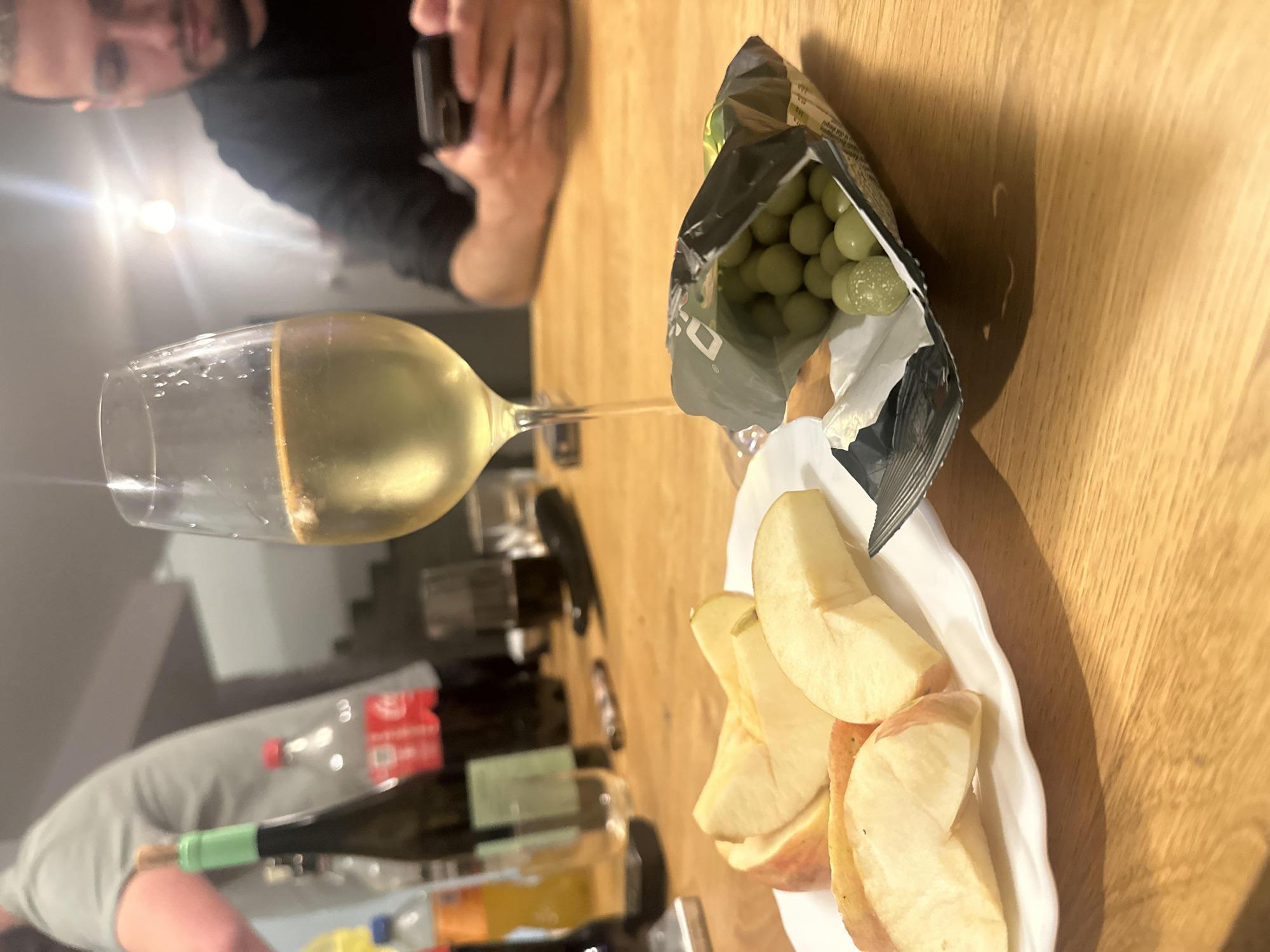 The image shows a table setting indoors. On the table are a wine glass filled with white wine, a bag of green candy or coated nuts, and a white plate with slices of apple. In the background, partially visible, there are several bottles, including what appears to be a wine bottle with a green label and a Coca-Cola bottle. Two people are partially visible. One person in the upper left is looking down at the scene and using a smartphone. Another person is visible at the bottom left of the image. The table appears to be made of wood, suggesting a home or restaurant setting. The location is not determinable from the image alone.