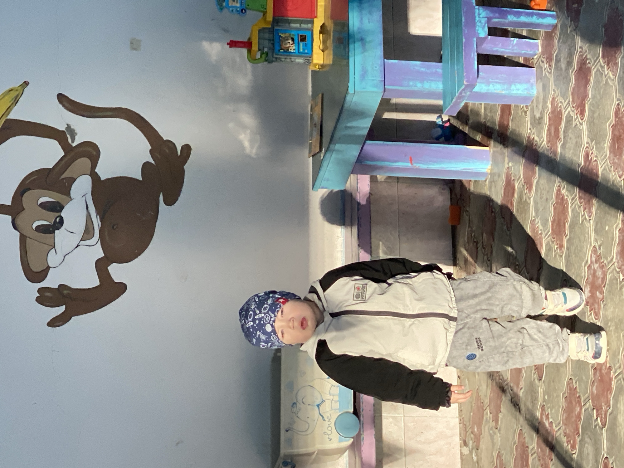 This indoor photograph, presented here in its upright orientation, captures a young child within a brightly lit room that appears to be a play area. On the left wall, a large, colorful mural of a cartoon monkey holding a yellow banana adds a playful element to the space. In the background, to the right, is a multi-tiered children's play structure or shelving unit, painted in vibrant shades of blue and purple. A small toy structure, featuring a red top, a yellow section, and a blue window with a toy figure inside, rests on top of this unit. The floor is covered in patterned tiles, displaying an intricate, repeating design in earthy tones.
The main subject is a toddler, standing and looking towards the camera with a neutral, slightly curious expression. The child is dressed in a blue beanie hat with white patterns, a two-tone white and black padded jacket, and grey sweatpants. Visible text on the sweatpants reads "STAY FRES" alongside a sports logo. The child also wears white sneakers with subtle colorful accents. To the child's left, a light blue container or toy box is visible, adorned with an elephant drawing and the word "Love."
Strong, distinct shadows are cast across the tiled floor and the lower portion of the wall by the child and the background structures, indicating a powerful light source, likely natural sunlight streaming in from a window. This suggests it is daytime, possibly mid-morning or late afternoon. The scene conveys a bright, cheerful, and child-friendly atmosphere, with no specific activity taking place other than the child's presence in the room.