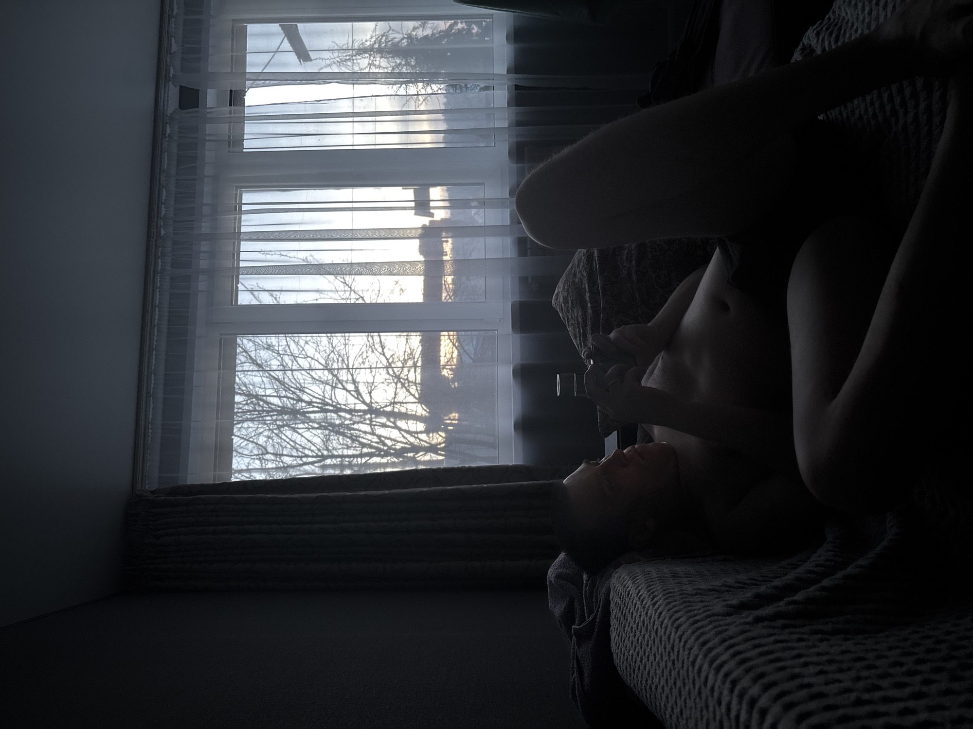 The image displays an indoor scene within a residence in Chaikovsky, Russia. A male individual, appearing shirtless and wearing dark lower garments, reclines on a couch or bed, positioned left of center. He holds a smartphone with both hands, focusing his gaze on its screen. A faint metallic chain is visible around his neck. The couch is partially covered by a textured, light-colored blanket on the left, while its upholstery underneath exhibits a dark pattern. A clear glass containing liquid rests on a surface next to the subject's head. The background is dominated by a large, multi-pane window dressed with translucent white sheer curtains and darker, pleated curtains drawn to the left. Outside the window, bare deciduous trees and a coniferous tree are visible, with distant building silhouettes against a sky displaying the low glow of either sunrise or sunset. The interior is dimly lit, relying primarily on ambient light from the window.