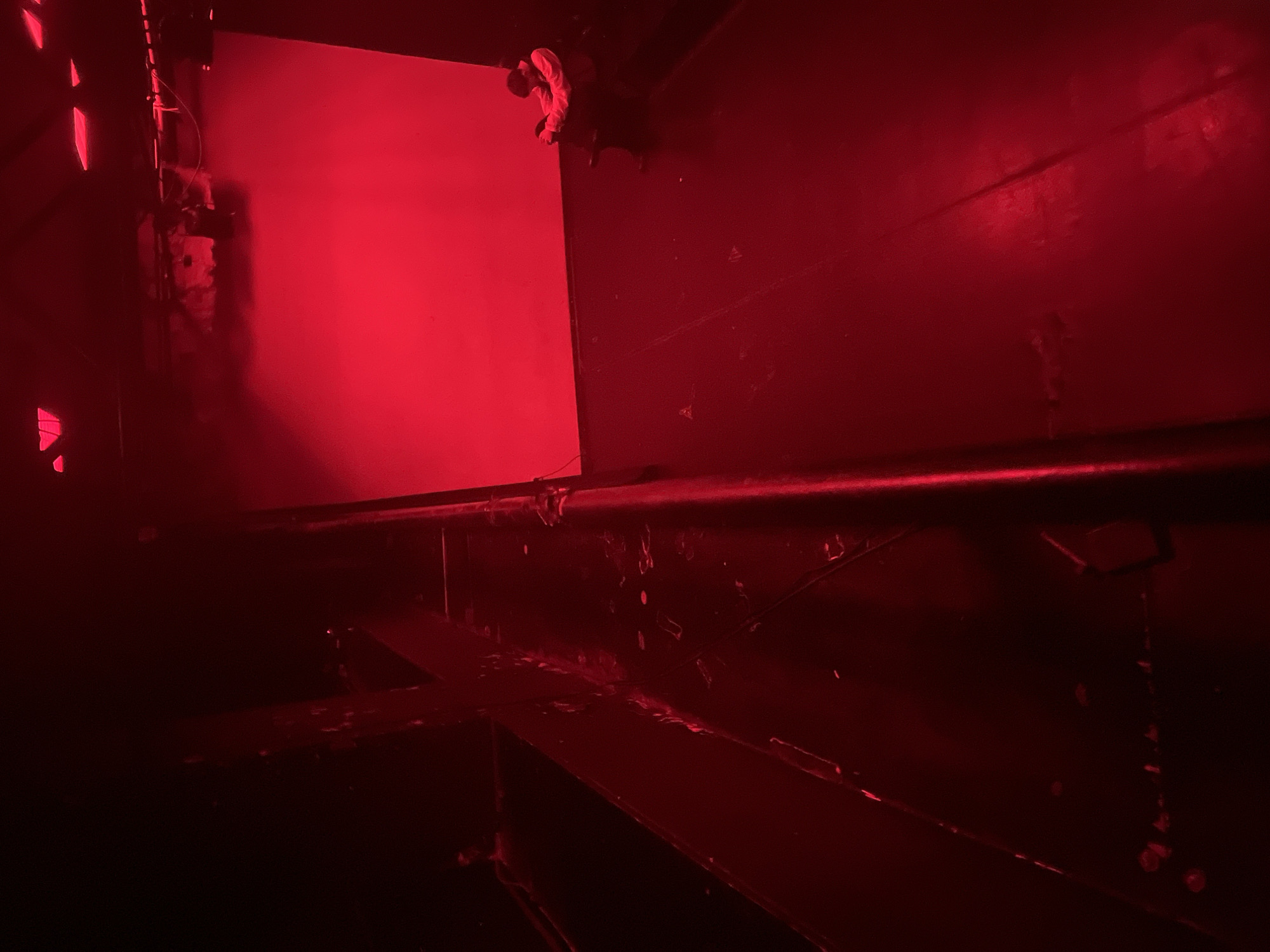 This media file captures an interior scene in Sofia, Bulgaria, dramatically illuminated by a pervasive, intense red light. The deep crimson hues dominate the frame, creating high contrast between intensely lit areas and dark, shadowy sections.
The main subject is a human figure positioned in the upper right quadrant of the image. The individual appears to be sitting or kneeling on a high, dark ledge or beam, looking down or across into the space. They are wearing light-colored clothing, likely a long-sleeved top, which stands out against the dark, red-tinged background. Their activity is not clearly discernible, but they seem to be focused on something in the immediate vicinity.
To the left, a large, rectangular panel or screen glows brightly with the same strong red light, acting as a prominent feature and possibly a primary light source within the space. Several dark, structural elements crisscross the scene, suggesting an industrial or constructed environment. A thick, dark horizontal bar or pipe runs across the mid-ground of the image, with other angular beams and supports visible below it, all reflecting glints of the red light. The overall texture appears metallic and somewhat rough.
The artificial red lighting imbues the entire scene with a stark, mysterious, and potentially industrial or artistic atmosphere. Details in the heavily shadowed areas are largely obscured. There is no visible text in the image. The time of day is not discernible due to the specialized lighting.