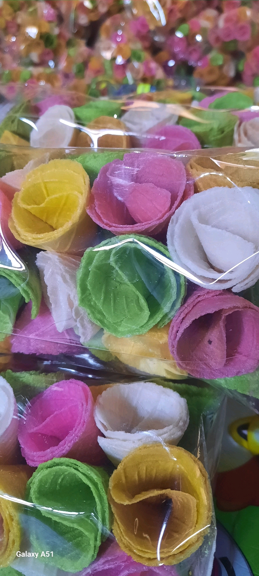 A close-up, slightly blurred vertical shot shows rows of colorful, rose-shaped cookies packaged in clear plastic. The cookies are arranged in concentric circles and come in various pastel shades including white, yellow, pink, green, and purple. The cookies appear to be made of a thin, crispy wafer. The background is also filled with more of these cookies, out of focus. A watermark "Galaxy A51" is visible in the bottom left corner. The image depicts a product likely sold in a market or store in Jatitengah, Indonesia.