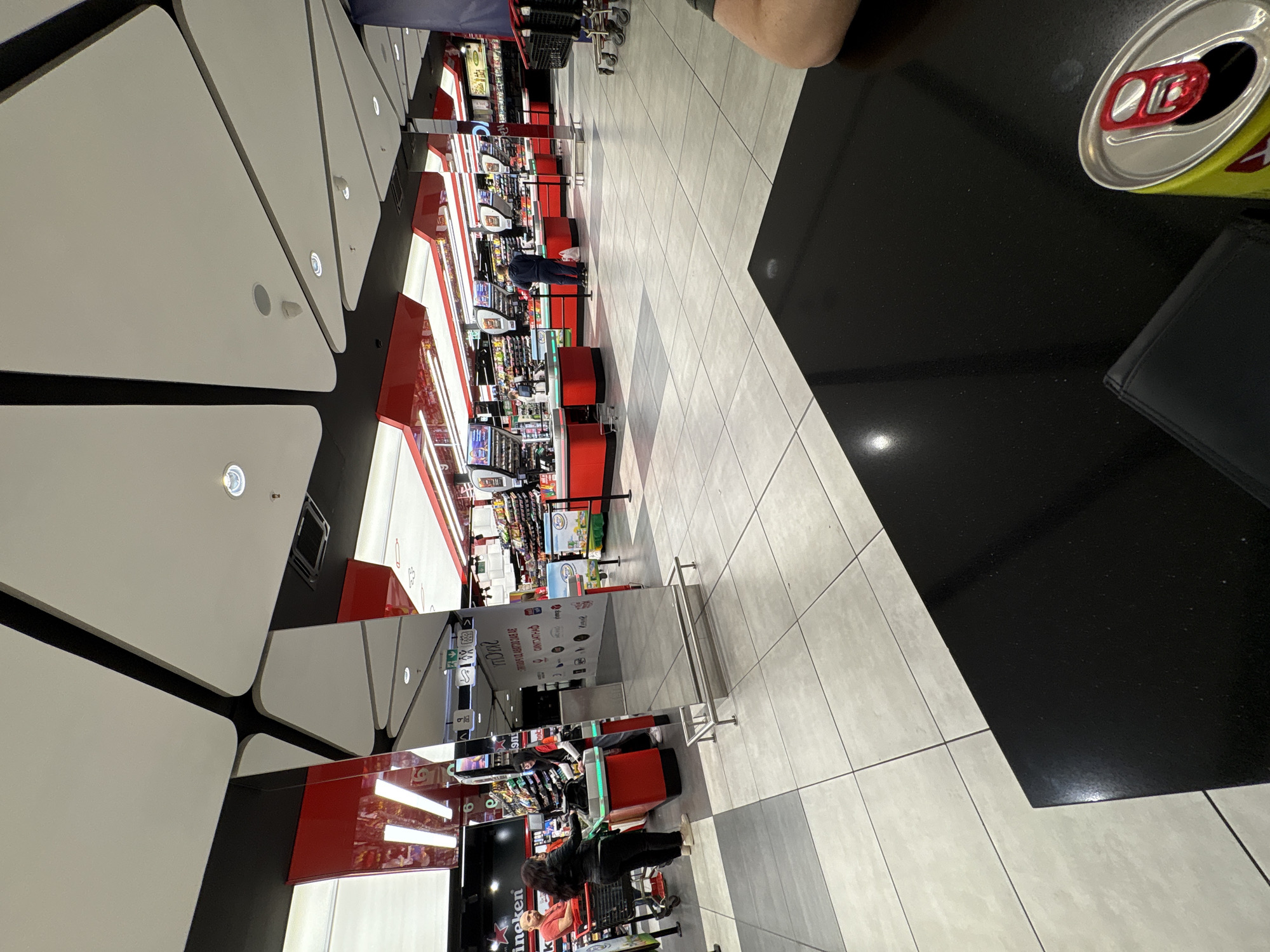 The image captures an overhead perspective inside a retail establishment, likely a supermarket or hypermarket, in Sofia, Bulgaria. The location is discernible by the Cyrillic script on displayed signage. Multiple checkout lanes, characterized by red and white color schemes, stretch into the distance. The area appears active, with both employees and customers engaging in transactions.
On a black, presumably granite, table in the foreground sits an open can of "Ariana" beer, adjacent to a dark wallet. A portion of a person's arm and shoulder is also visible, indicating someone is sitting at the table.
The ceiling has a modern design consisting of white triangular panels trimmed with black. The floor is tiled in light gray, with darker gray inlays, to define the store layout.