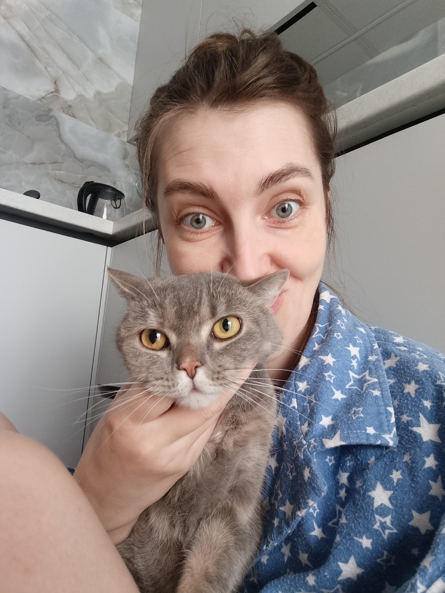 A selfie shows a woman indoors, holding a gray cat with yellow eyes. The woman has fair skin, light-colored eyes, and her brown hair is styled in a bun. She is wearing a blue star-patterned pajama top. The cat's face is centered, its nose close to her mouth. A hand is visible, holding the cat under its chin. In the background, there is a white cabinet and a wall with a marble-like pattern. A black electric kettle is visible on the cabinet. The scene appears to be in a residential kitchen. There is no evidence to confirm the location is Chaikovsky, Russia.