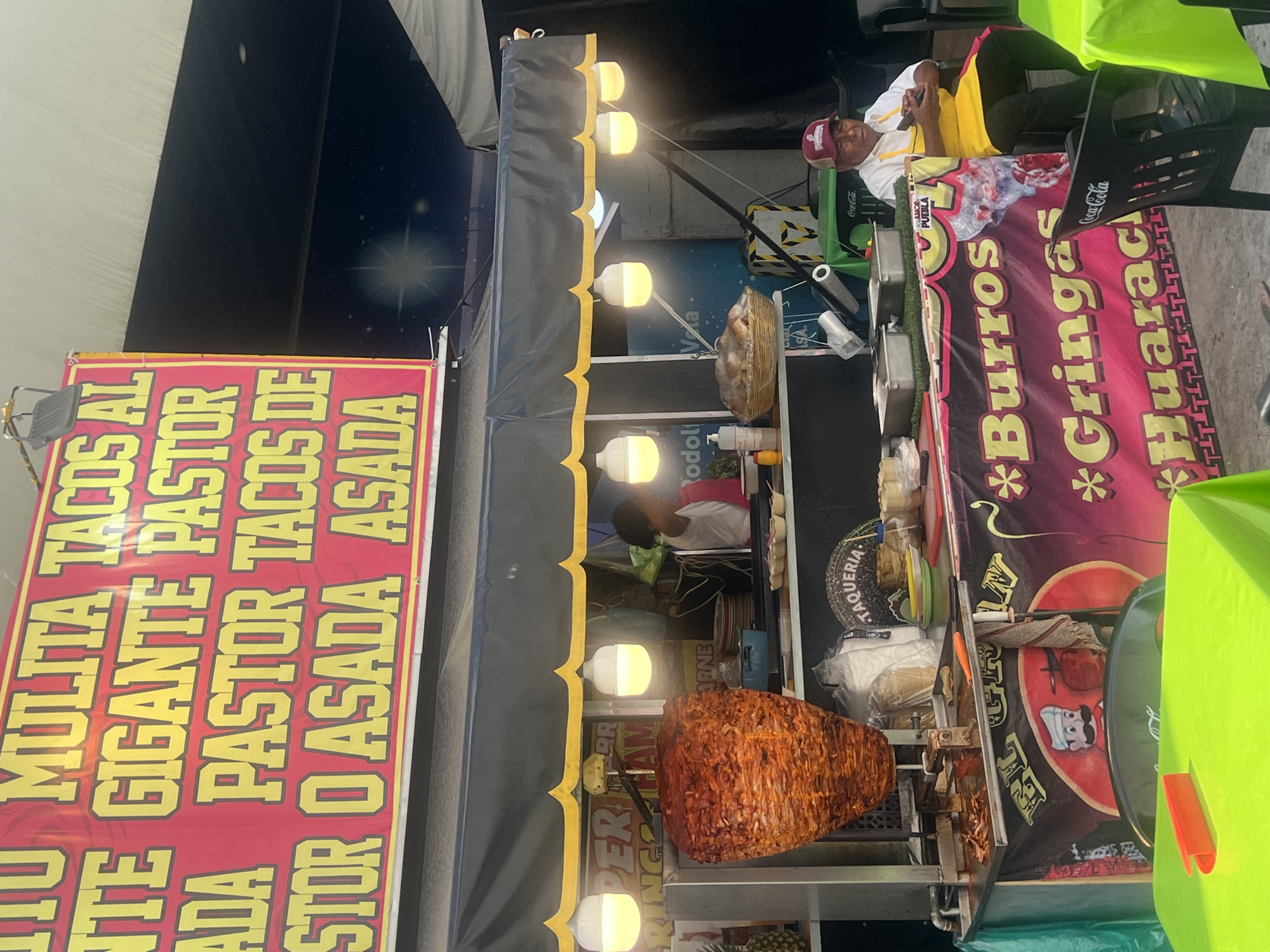 A bustling street scene captures a food vendor in Unknown, Unknown. A man in a red baseball cap and yellow shirt sits on a green chair beside a Coca-Cola branded banner featuring the words "Burros", "Gringas" and "Huarach". Adjacent to the banner is a large, red and yellow sign advertising "Mulita", "Tacos Al", "Gigante Pastor", and "Tacos De Asada". A revolving grill showcases a large portion of meat for tacos. The food stand is adorned with a series of light bulbs under a black awning. The street is filled with activity, reflecting the vibrant urban environment.