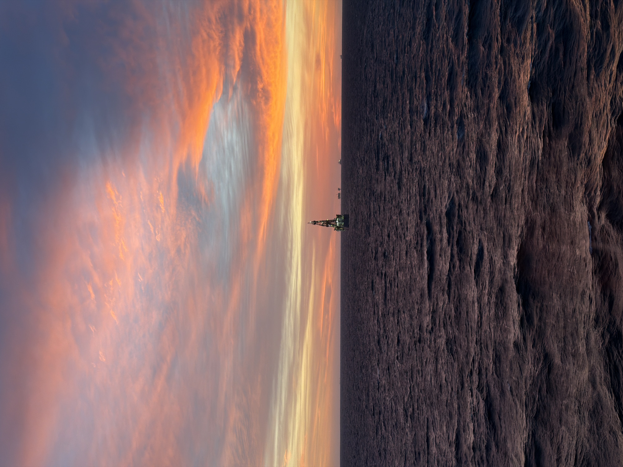 The image displays a dramatic sunrise or sunset over a vast expanse of water, likely an ocean or sea, with an offshore industrial structure prominent on the horizon.
The sky, occupying the upper half of the frame, is a spectacle of color. It transitions from deep blues and purples at the top, through soft pinks and light blues, to intense oranges, yellows, and fiery reds directly above the horizon where the sun's low angle illuminates widespread clouds. These clouds are streaky and elongated, beautifully catching the warm light.
Below the distinct horizon line, the water appears dark and deep, almost black or a very dark indigo, reflecting little of the sky's brilliance. The surface exhibits subtle texture, suggesting gentle waves or currents.
An offshore oil rig or similar platform is centrally positioned on the horizon. Its dark silhouette is punctuated by numerous bright yellow and white lights, indicating it is operational against the dimming or brightening sky. Several smaller, indistinct structures are visible further along the distant horizon.
The time of day is either sunrise or sunset, characterized by the vibrant, warm hues in the sky and the low light conditions that necessitate the rig's illumination. The atmosphere is serene yet industrial. There are no people visible, nor is there any discernible text.