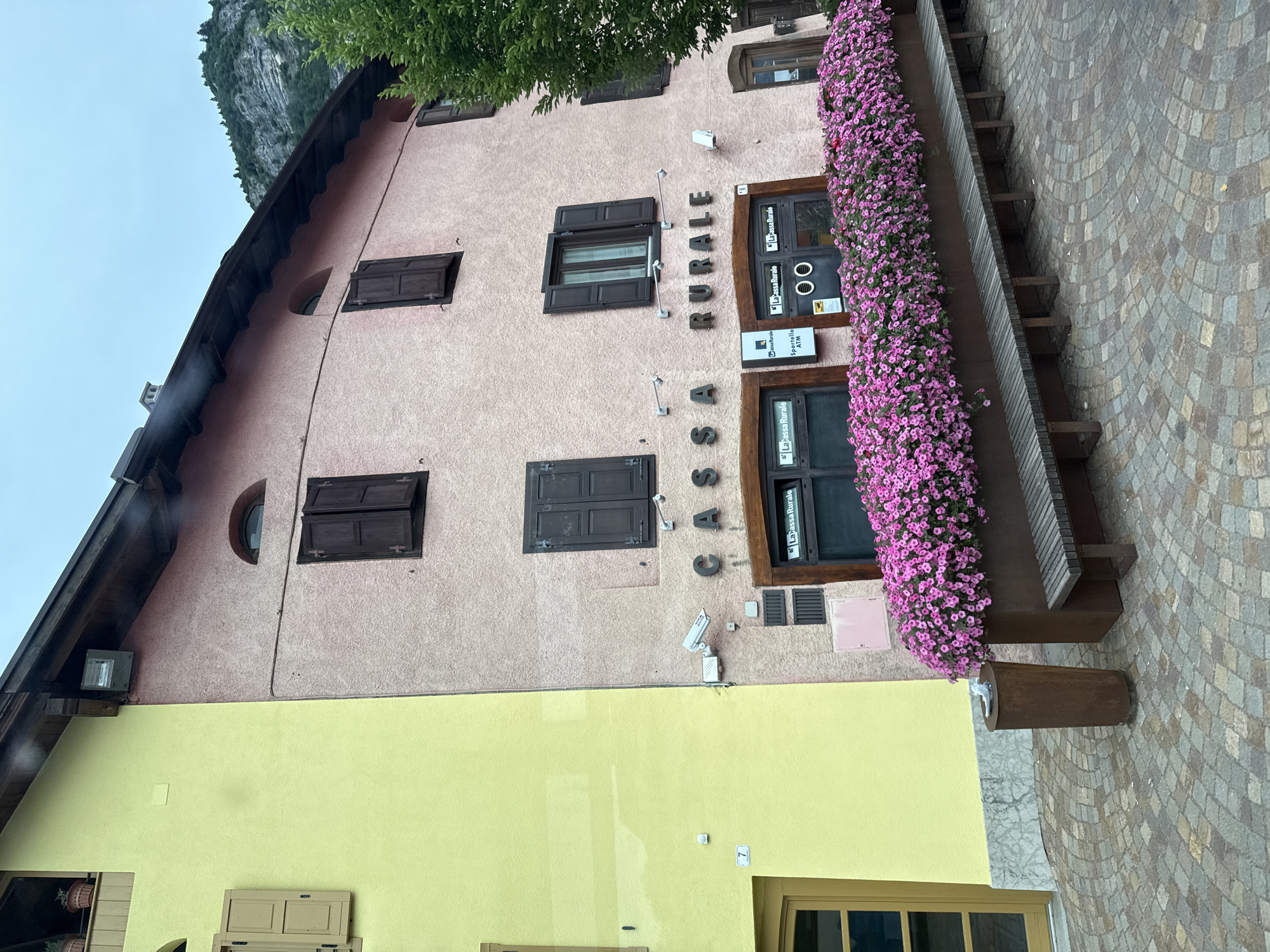 The image shows the exterior of a building in Fai della Paganella, Italy. The building has a pink facade with several windows and shutters. Above the entrance, large metal letters spell out "CASSA RURALE." Below this, there is a storefront with a window displaying "La Cassa Rurale Sportello ATM." A long planter filled with vibrant pink flowers runs along the front of the building, with a wooden bench placed in front of it. A cylindrical trash can is located to the left of the planter. The pavement in front of the building is paved with cobblestones arranged in a circular pattern. A green tree is visible on the right side of the image, and a mountain is in the background. The sky is overcast.
