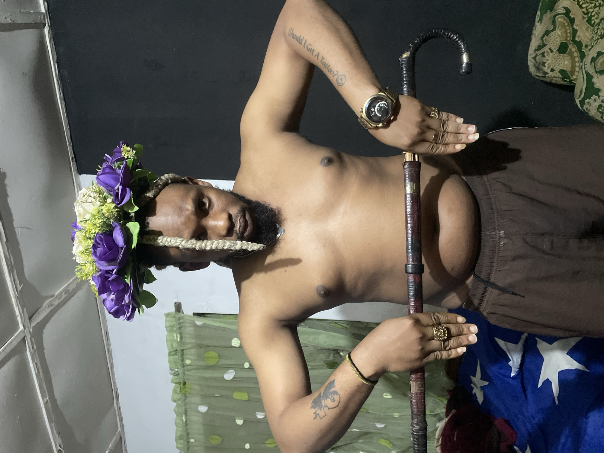 A shirtless man with a beard and a flower crown is holding a cane in an indoor setting. The man has tattoos on his left arm and a watch on his right wrist. He is wearing brown shorts. The background includes a green curtain with white polka dots on the left and a black wall on the right. There are also some folded cloths with white stars on a blue background on the left, and a patterned cushion on the right. The lighting suggests an artificial light source. The scene does not contain any elements that specifically identify its location within Jos, Nigeria.