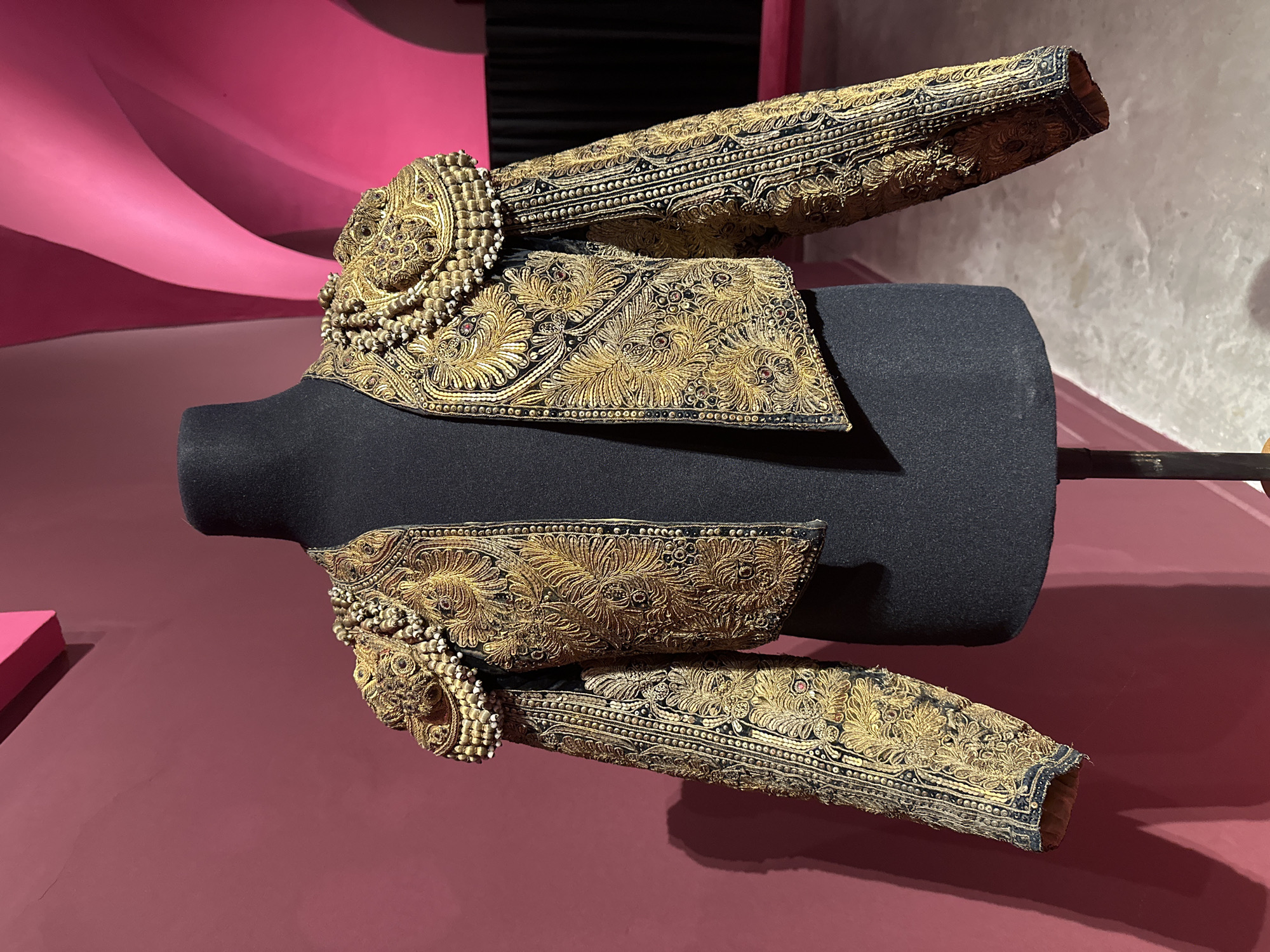 A detailed, ornate gold and black garment, possibly a matador's jacket, is displayed on a dark gray torso form on a burgundy-colored surface. The jacket features elaborate gold embroidery and embellishments, including beadwork around the shoulders. The background includes a pink curved structure and a section of black, possibly fabric or wall covering, providing contrasting colors. The location is likely in Buenos Aires, Argentina, given the cultural significance of matador garments in the region.