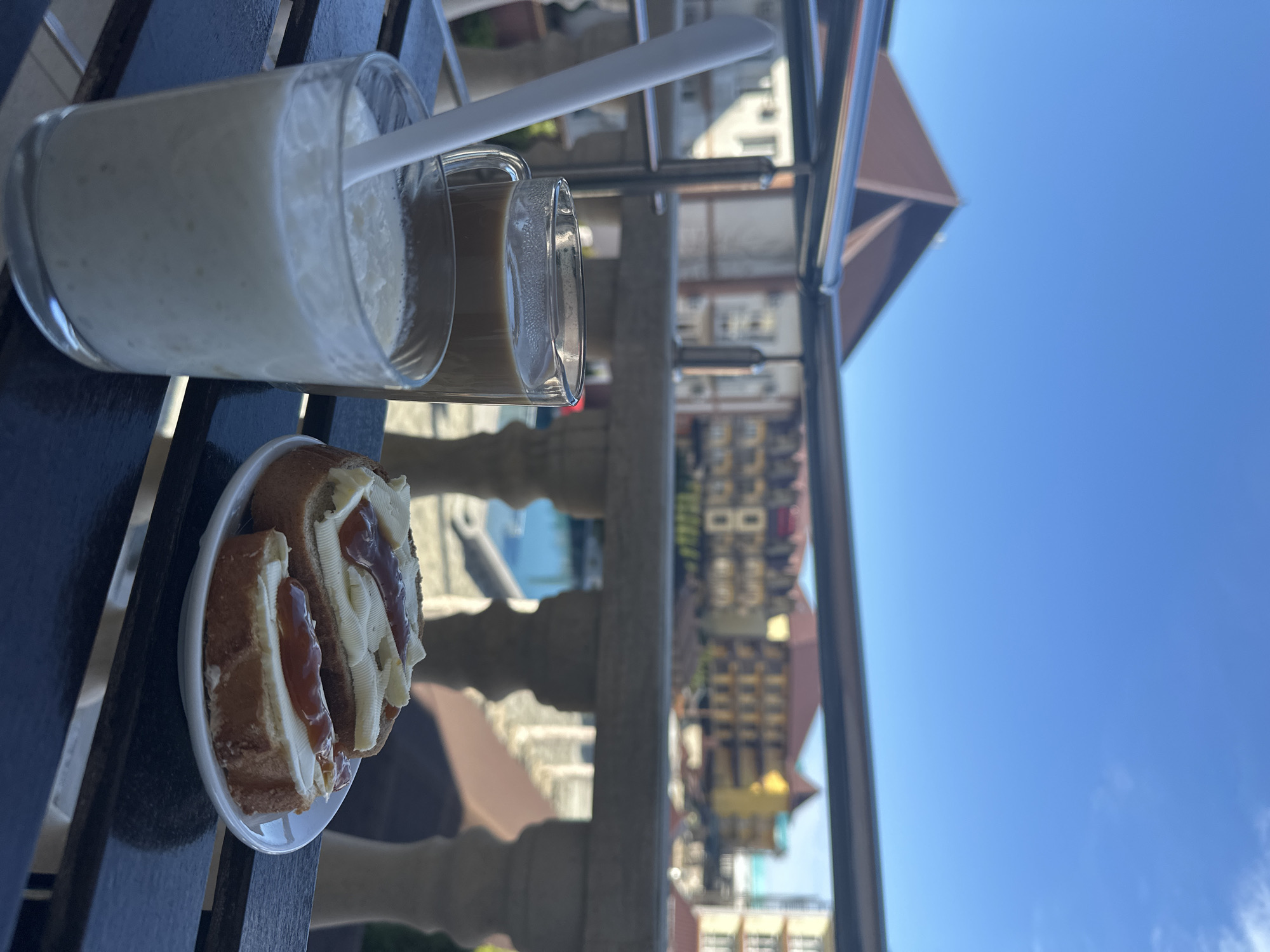 A plate of sandwiches and two glasses of drinks sit on a dark surface on a balcony. One glass contains a white, frothy drink, while the other holds a brown liquid. A white spoon rests across the top of both glasses. The sandwiches appear to be filled with cream and a reddish-brown spread. In the background, a railing obscures the view of a city with buildings of various colors, with a clear blue sky above. The location is potentially Novomikhaylovsky, Russia, based on visual cues from the architecture and general aesthetic of the background.