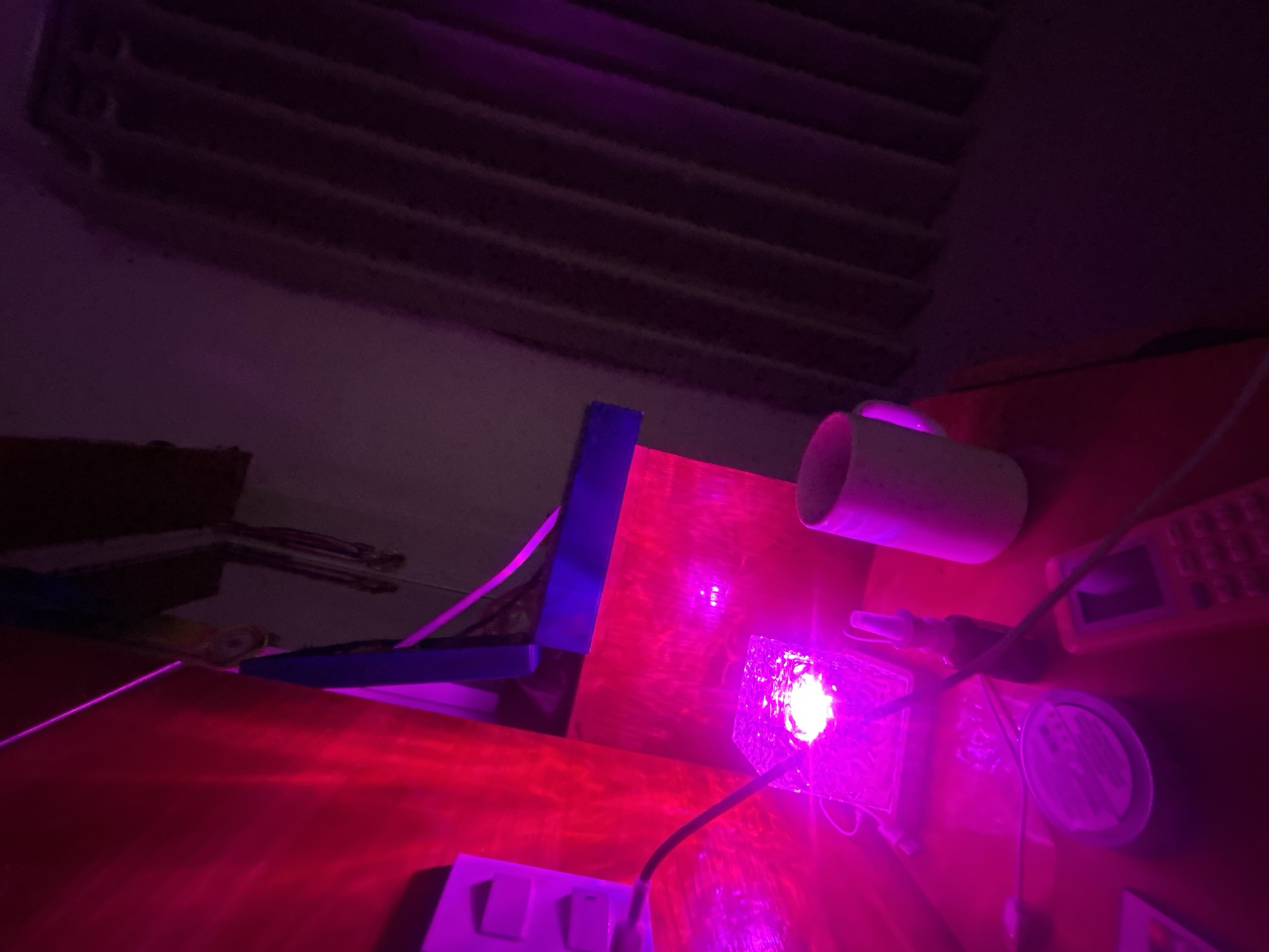 The image depicts a close-up, low-angle shot of a desk in what appears to be a domestic setting. The primary light source is a bright pink LED light illuminating a clear, cube-shaped object on the desk. The light reflects intensely off the object, creating a strong glow.
To the left of the cube, a white electrical outlet with two sockets is visible on a red wall. A black cable extends from the outlet and connects to the cube.
In the foreground, on the desk, several other objects are present:
* A small, dark-colored bottle with a white dropper cap, possibly a spray bottle.
* A white mug, positioned to the right of the cube.
* A circular, flat object, possibly a coaster or a container lid.
* A pink electronic device, resembling a small phone or calculator, is positioned in the lower right quadrant.
The background is dominated by dark, vertical blinds or curtains. The overall lighting is dim and saturated with pink and purple hues, suggesting either a deliberate lighting setup or low ambient light conditions.
The scene is devoid of any individuals or clear signs of human activity. The precise location within Nha Trang Ward, Vietnam, cannot be determined from the visual information provided.