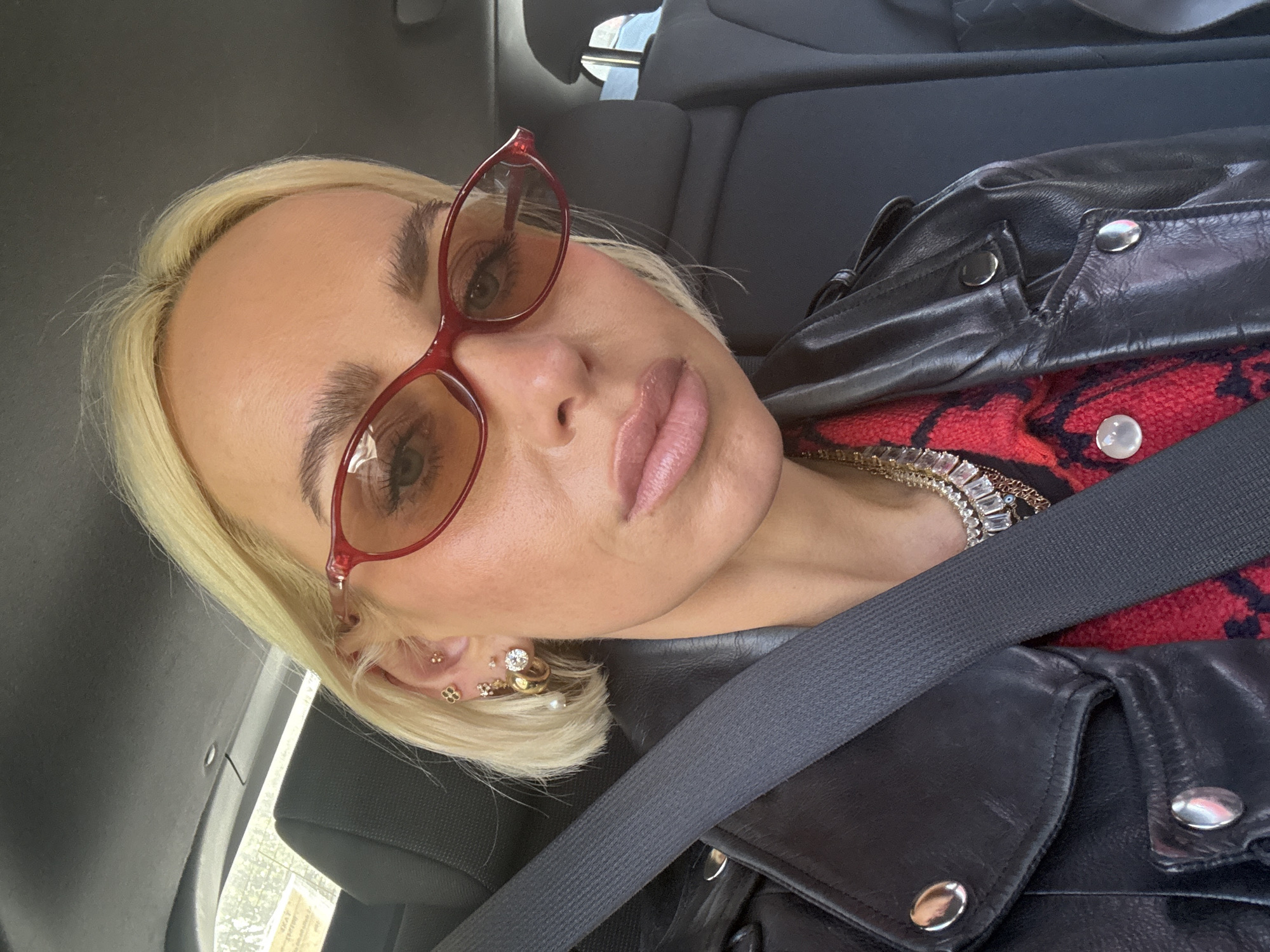A blonde-haired woman wearing red sunglasses and a black leather jacket with silver buttons is sitting in a car, secured by a gray seatbelt. She has multiple earrings in her left ear and wears a red and black patterned top with a layered necklace visible. The car appears to be parked, and the location is unspecified, but given the prompt, it is understood to be within the city of Milan, Italy. The car has black seats and a dark gray interior. The woman is looking directly at the camera.