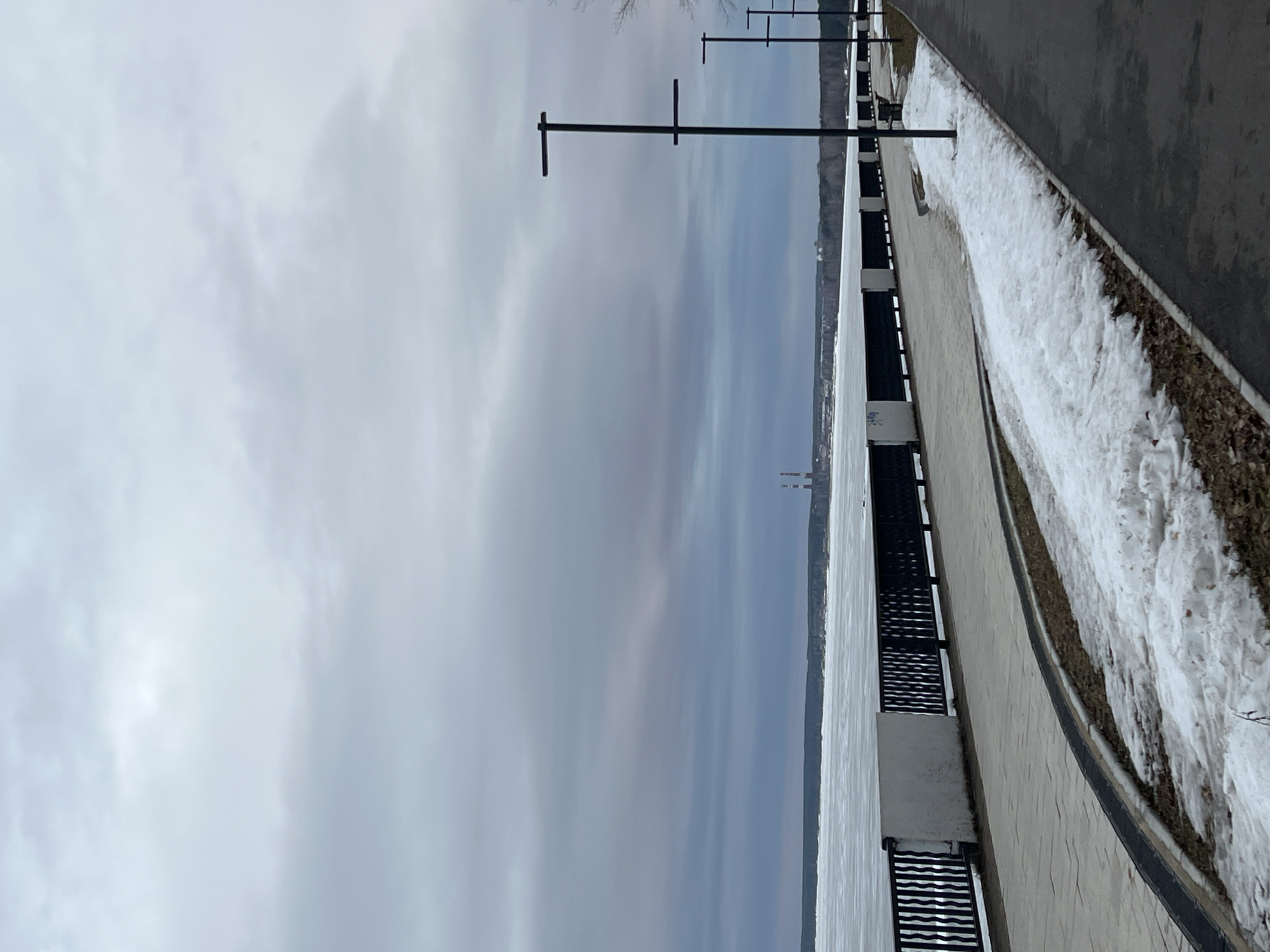 The image depicts a view of a frozen body of water, likely the Kama River, near the city of Chaikovsky, Russia. The scene is shot from a slightly elevated position along a paved embankment or promenade, which is lined with a black metal railing and white concrete posts. There are light poles spaced evenly along the embankment. A snow-covered area runs parallel to the pavement. The river is largely covered with ice, and in the distance, what appear to be industrial chimneys can be seen. The sky is overcast and grey, indicating cold, possibly winter weather conditions.