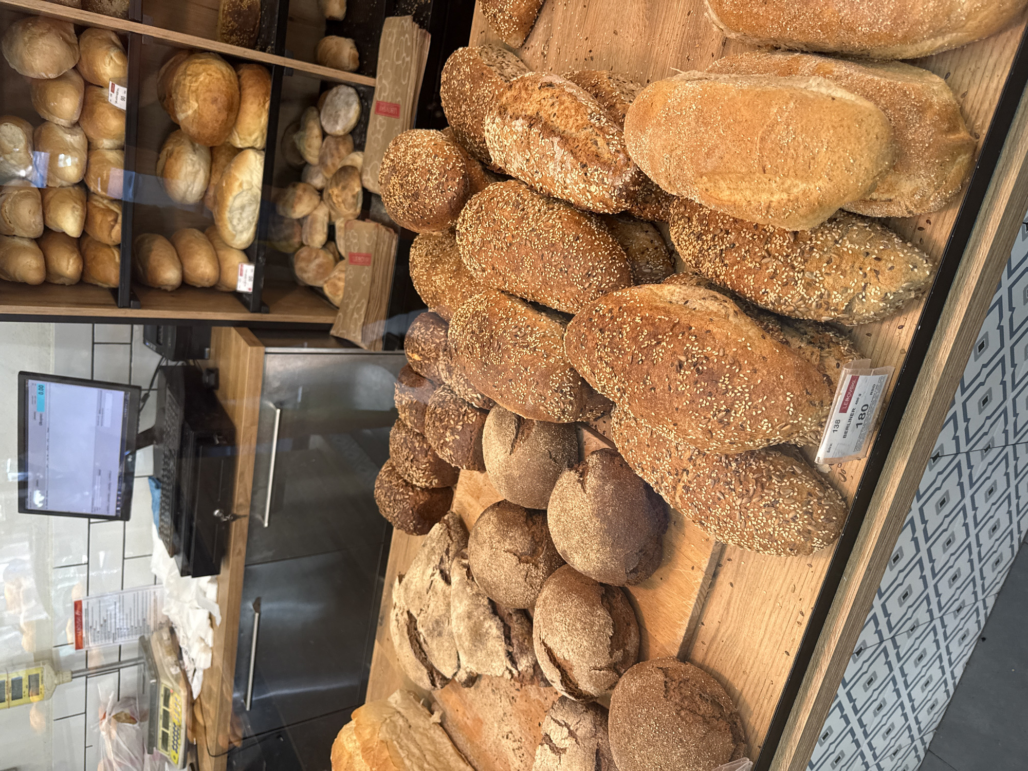 This is a medium shot, eye-level, indoor photograph of a bakery display in Belgrade, Serbia. The display features a variety of bread loaves and rolls arranged on a wooden surface, with a glass partition separating the viewer from the baked goods. In the background, a shelf displays more bread. A POS system, including a monitor, keyboard, and cash drawer, is visible on a counter to the left. A digital scale is also present on the counter. The overall lighting is bright and even. There are no people in the image. The scene is clean and organized, suggesting a retail environment.