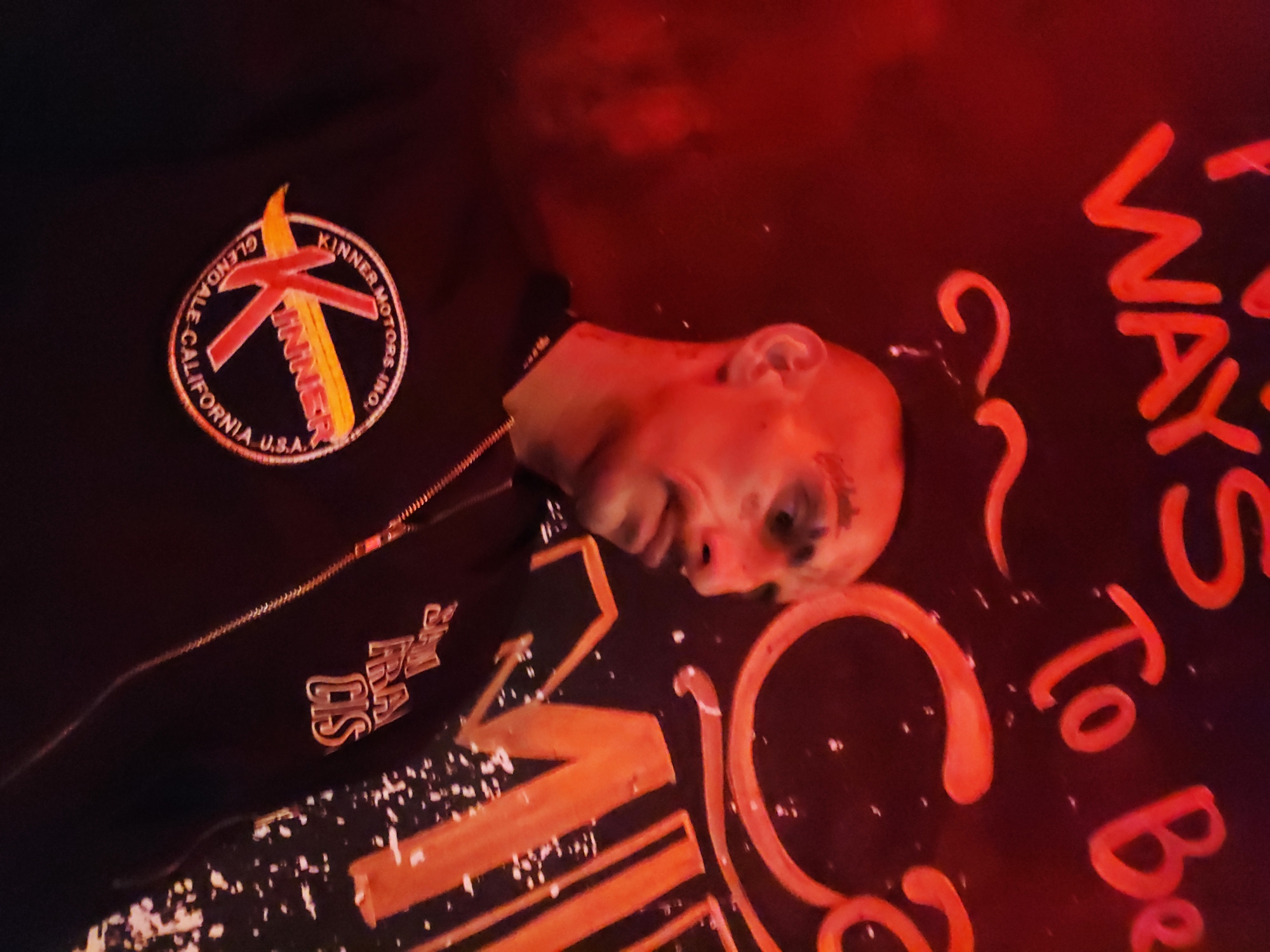 The image shows a man wearing a black jacket with "KINNER" embroidered on the left breast and "SAN FRA DIS" on the lower left. The jacket features a gold zipper. The man's face is illuminated with red light. The backdrop appears to be a sign with writing on it, including the words "WAYS To Be". The environment is dark and appears to be indoors. There is no specific reference to Nha Trang Ward, Vietnam in the image.