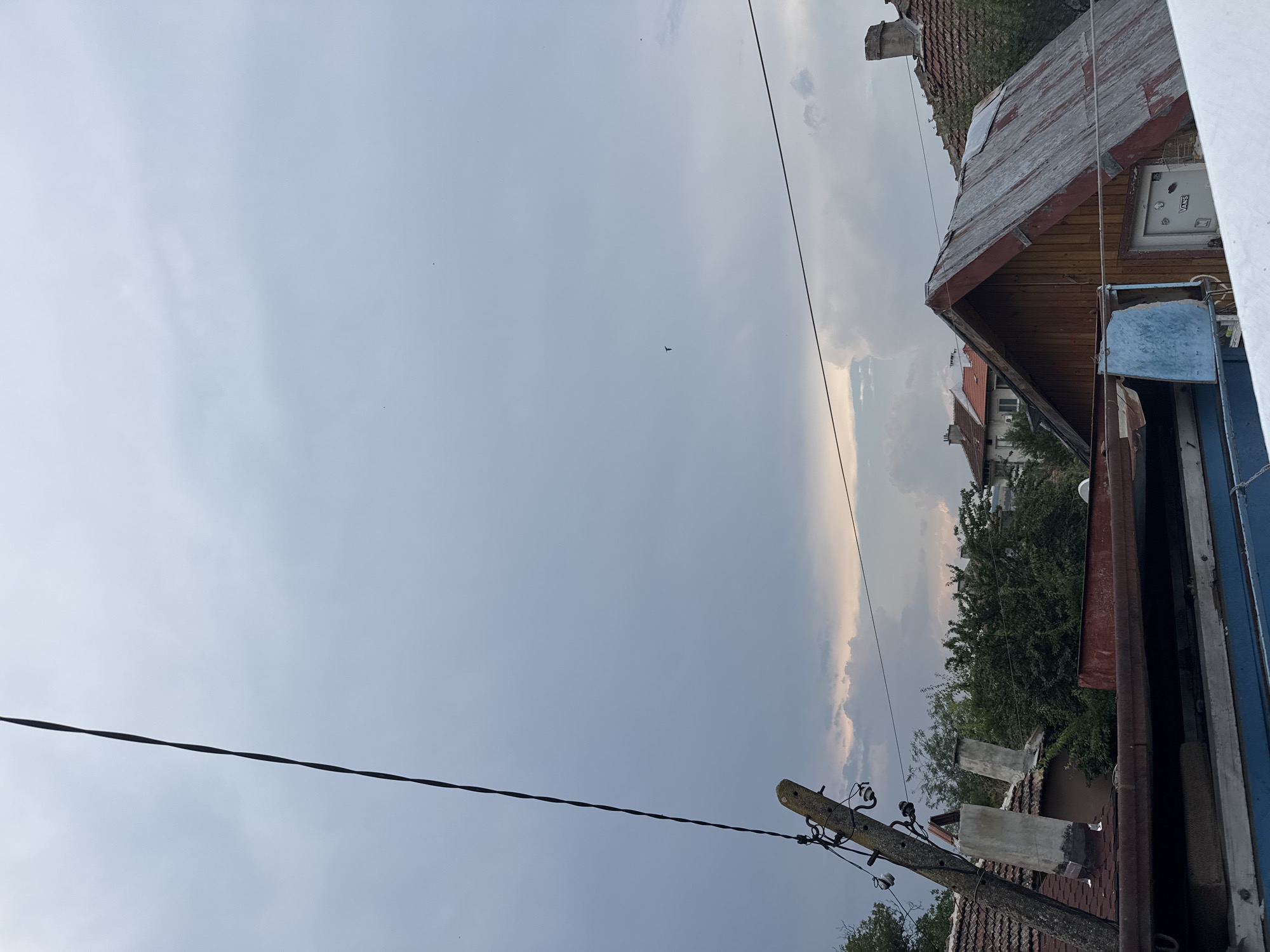 The image depicts a view from an elevated vantage point looking towards a residential area in the city of Sredets, Bulgaria. The sky is overcast with gray clouds, suggesting a potential for rain or a recent storm, with a sliver of lighter sky visible towards the horizon. A power line runs diagonally across the frame from the top left corner. In the foreground, there are several structures with corrugated metal roofs and wooden elements. To the left, a weathered utility pole with insulators stands prominently. Behind it, the roofs of houses with red tile and corrugated metal are visible, interspersed with lush green trees. A small, dark bird is seen flying in the sky. The overall impression is a somewhat utilitarian and weathered urban landscape under a moody sky.