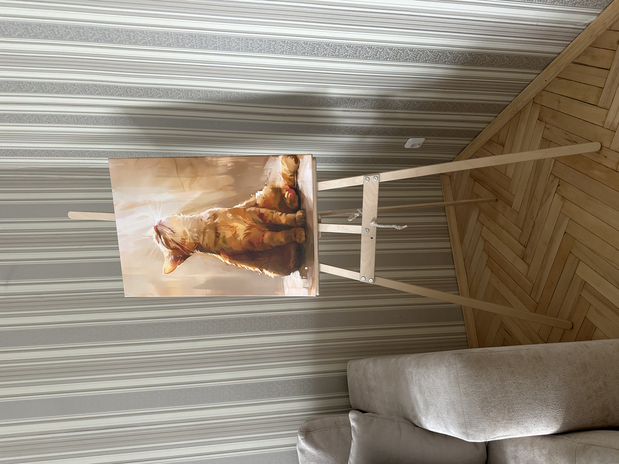 The image depicts an indoor scene, featuring a painting of an orange cat on an easel positioned in the corner of a room. The painting is illuminated by natural light, creating a warm glow on the feline subject. To the left of the easel, a portion of a light-colored couch is visible. The room is decorated with striped wallpaper in shades of grey and beige. The floor is made of light-colored wooden parquet arranged in a herringbone pattern. An electrical outlet is visible on the wall behind the easel. There are no people present in the image, and no specific location in Minsk, Belarus, can be identified from the visual cues provided. The overall atmosphere is calm and domestic.