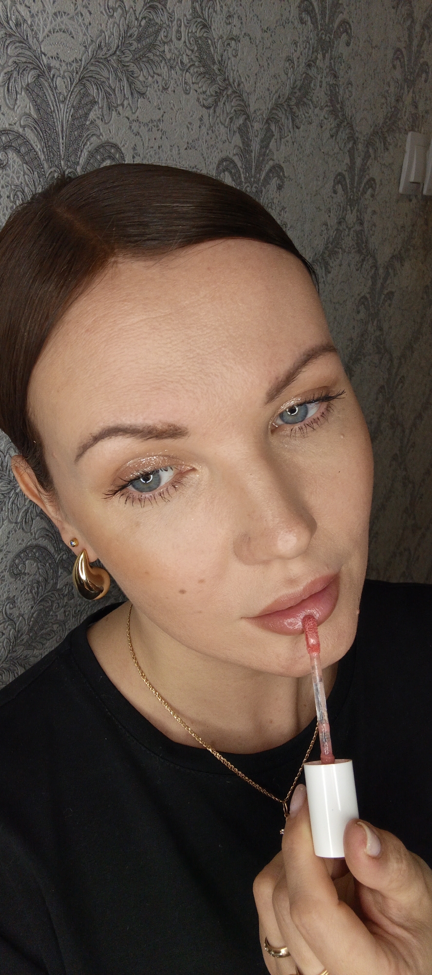 A person is shown in a close-up, indoor shot applying lip gloss. The person has brown hair pulled back, blue eyes, and is wearing gold earrings and a gold necklace. They are holding a lip gloss applicator to their lips, and the tube of lip gloss is visible in their hand. The person is wearing a black t-shirt. The background is a wall with a grey damask pattern. There is no evidence in the image to determine the location in the city of Chaikovsky, Russia.
