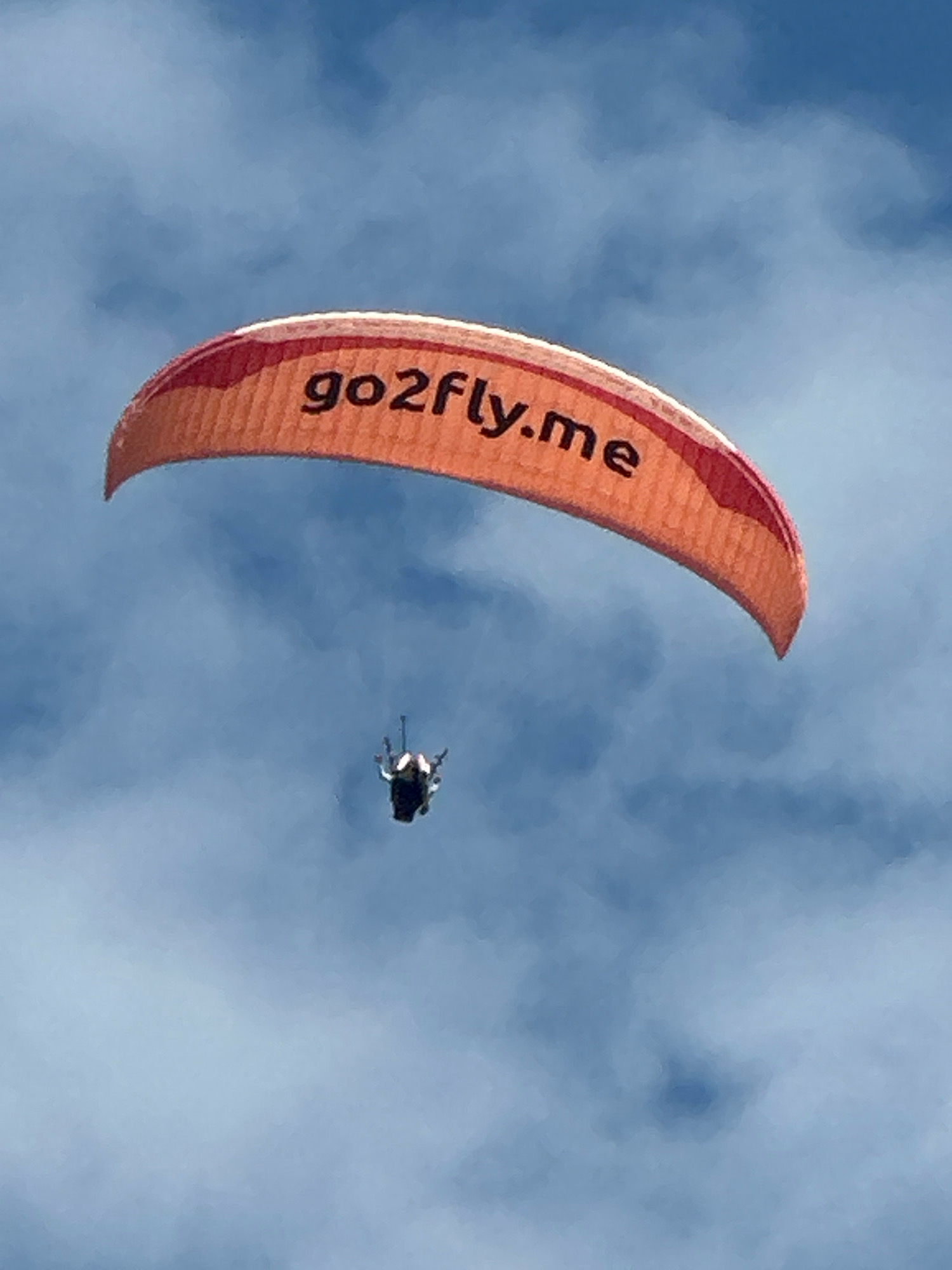 An orange paraglider with the text "go2fly.me" printed in black is visible against a blue sky with scattered clouds. Below the paraglider, a person is suspended in a harness. The paraglider is positioned in the upper portion of the image, while the person is in the lower middle. The scene appears to be an outdoor activity in a generally clear sky. There is no visible indication of the city of Boreti, Montenegro in this image.