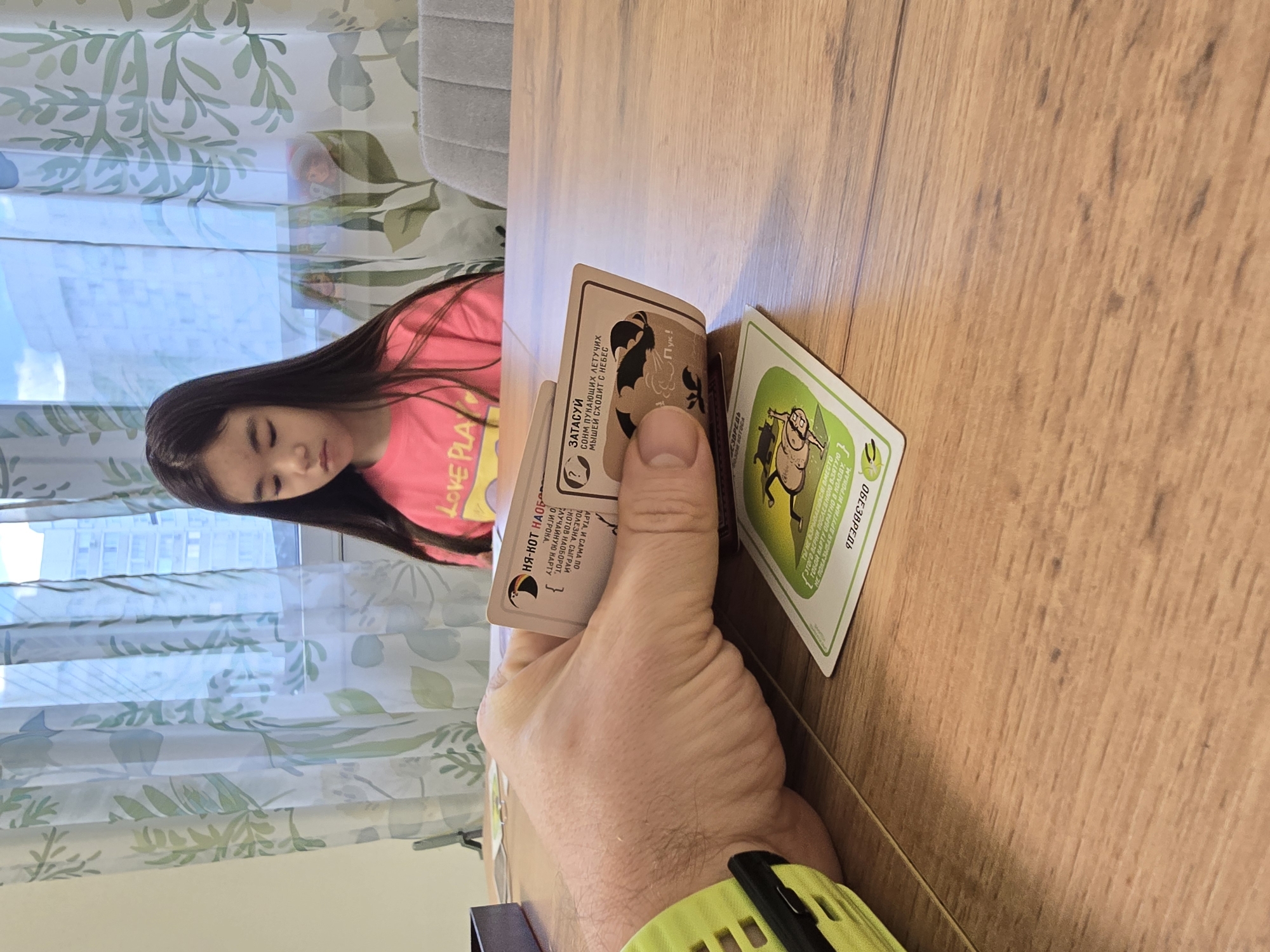 A photograph taken indoors depicts a hand holding a small stack of illustrated game cards over a wooden tabletop. The closest card to the foreground is cream-colored with Cyrillic text and a cartoon illustration. A green-rimmed card with a cartoon figure also lies on the table. The hand holding the cards is fair-skinned and wearing a lime green and black smartwatch.
In the blurred background, a young girl with long dark hair, wearing a pink shirt with yellow text, is visible, partially obscured by a window with light blue, patterned curtains. A gray chair back is also visible.
The photo was taken in Khimki, Russia.