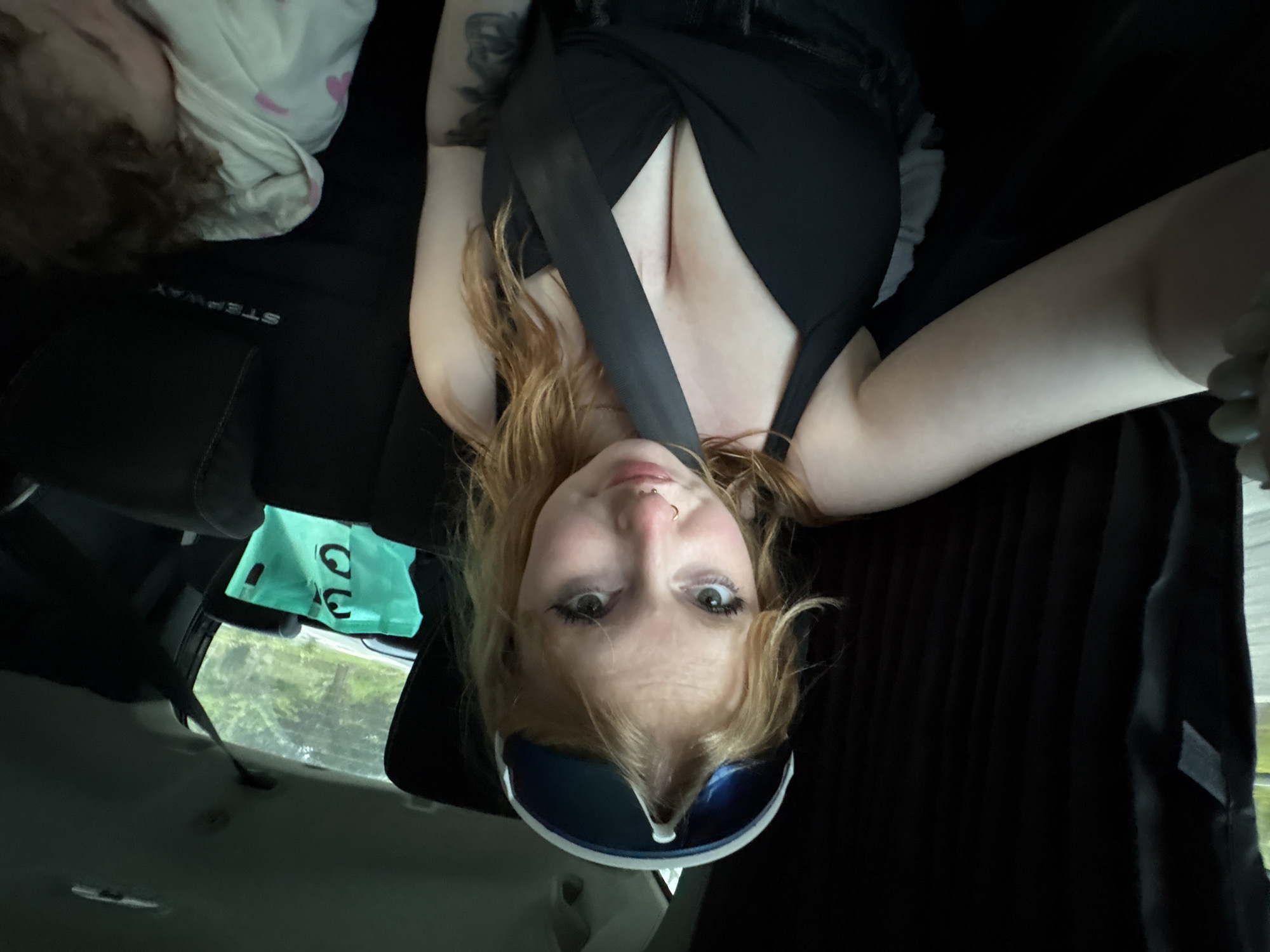 A low-angle, upside-down selfie shows a young woman in a car. She has fair skin, strawberry blonde hair, and a nose ring. She wears black sunglasses perched on her forehead, a black tank top, and a seatbelt. A black tattoo is visible on her left arm. In the background, the car's interior includes a black seat with the word "STEPWAY" printed on it, a window with a glimpse of green foliage, and a green plastic bag hanging from the ceiling.