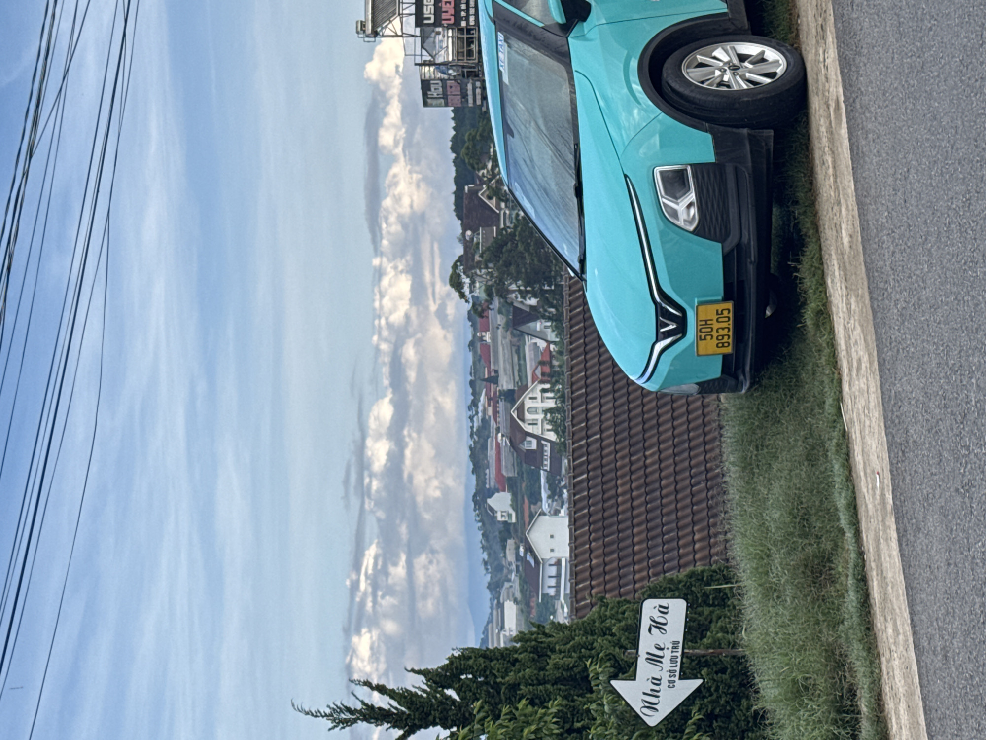 The image depicts a turquoise VinFast VF e34 electric car parked on the side of a road, overlooking a hillside town in Lâm Viên - Đà Lạt, Vietnam. The car's license plate reads "50H 893.05". A white directional signpost with Vietnamese text, likely indicating a location called "Nhà Me Hà" and "CƠ SỞ LƯU TRÚ", is positioned to the left of the car, partially obscured by a tall, dark green coniferous tree. The town in the background features a collection of houses with tiled roofs, set against a backdrop of hills and a cloudy sky. Overhead, power lines are visible against the sky. The foreground shows a grassy embankment and a paved road.