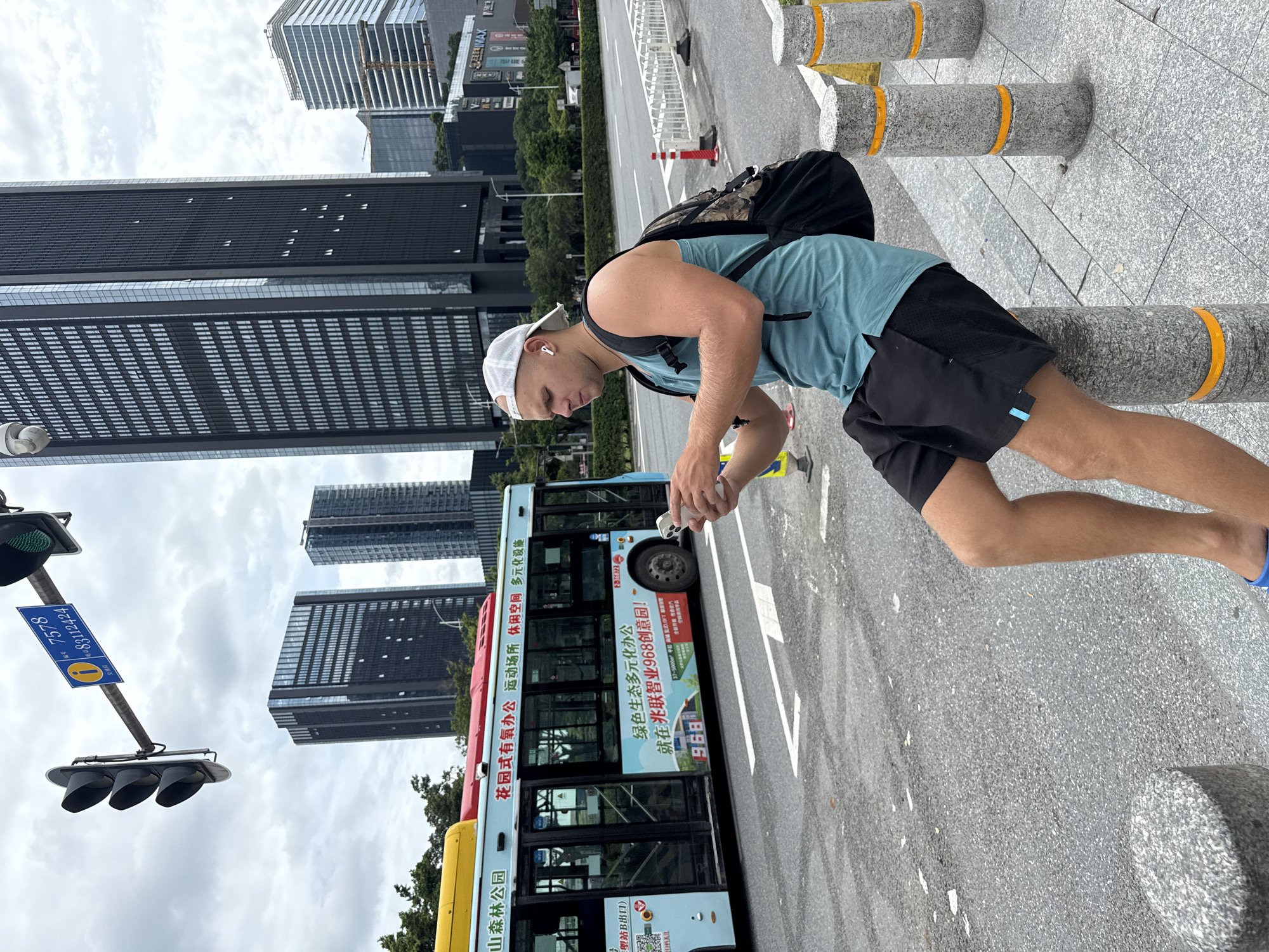 In Haizhu District, China, a young man wearing a teal tank top, black shorts, a white baseball cap, and white earbuds sits on a concrete bollard next to a street. He is looking down at his phone, which he holds in his hands. He has a black backpack on his shoulders. A light blue bus with Chinese text on its side is stopped in the background. Traffic lights and tall modern buildings are also visible. The sky is overcast.