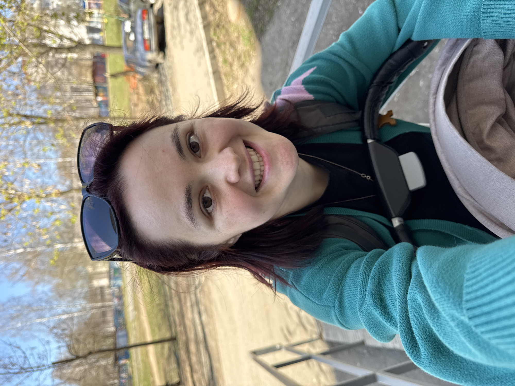 A medium shot shows a smiling woman holding a camera, in what appears to be a selfie, as she stands on a light gray tiled surface in Izhevsk, Russia. The background, out of focus, suggests an outdoor setting with trees and a road where a car is parked. The woman has dark hair, and is wearing a black top and a teal cardigan with a pink decorative patch on the shoulder. Sunglasses are perched on her head. Her left hand holds the camera up high, and it is positioned near the handle of a stroller, with a light tan-colored interior visible. The scene is in daylight, and there is natural lighting.