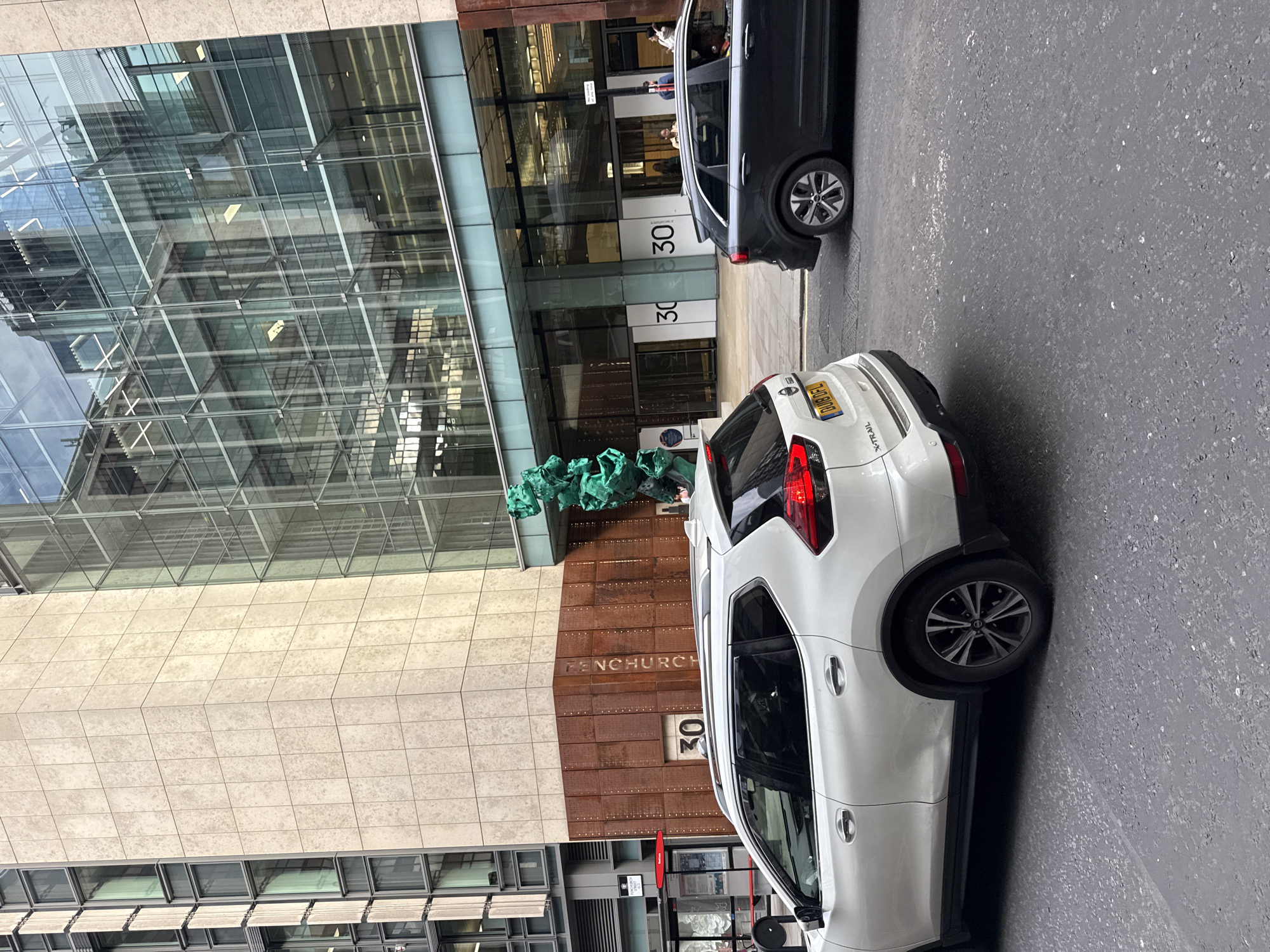 The image captures a street scene in the city of London, United Kingdom, likely in the financial district, given the modern architecture. In the foreground, a white SUV is parked on the right side of the street, with its rear facing the viewer. To its left, a dark-colored SUV is also parked. Behind the vehicles, a contemporary building dominates the scene. The building features a facade of light-colored stone on the left and a large glass-fronted section on the right, reflecting the surrounding environment.
Prominently displayed on the stone section of the building is a large number "30" and the text "FENCHURCH". A distinctive, bright green sculpture is positioned in front of the glass entrance of the building. Several people are visible near the entrance, some appearing to be entering or exiting the building. A bus stop shelter with advertisements is located to the left of the building, further indicating an urban environment. The street itself is paved with dark asphalt, and the lighting suggests it is daytime.