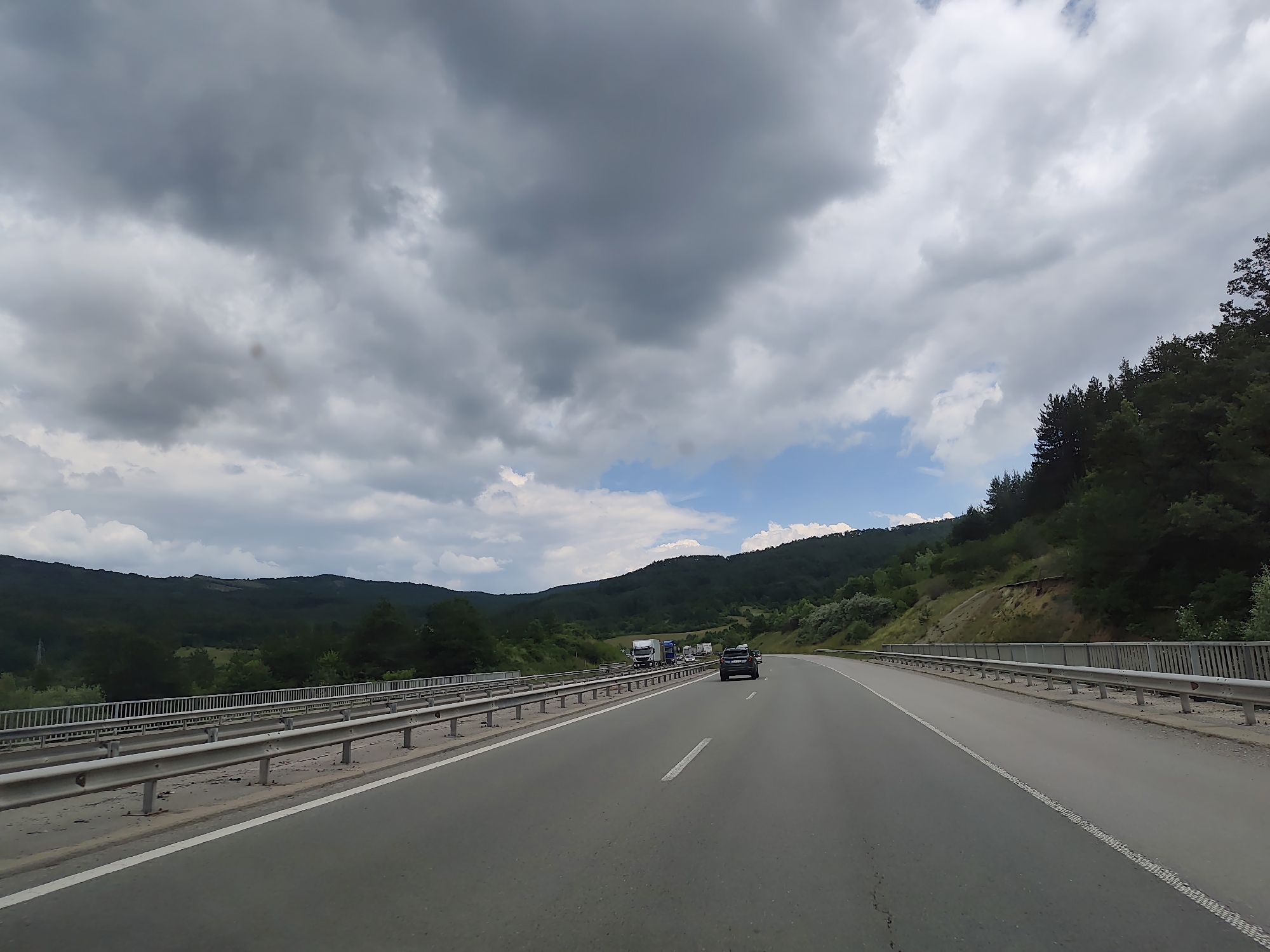 The image captures a view from a vehicle traveling on a highway, likely in or near Golemo Buchino, Bulgaria. The highway curves to the right, with a metal guardrail lining the left side. The road is asphalt, with a white dashed line marking the lanes. In the distance, on the left side of the highway, several large trucks are visible, suggesting a traffic slowdown or congestion. A black SUV is in the foreground, on the right lane of the highway, moving away from the camera. The surrounding environment is hilly and densely forested with various shades of green. The sky above is a mix of dark, heavy clouds and patches of bright blue sky with white clouds, indicating an overcast or partly cloudy day. The lighting suggests it is daytime.