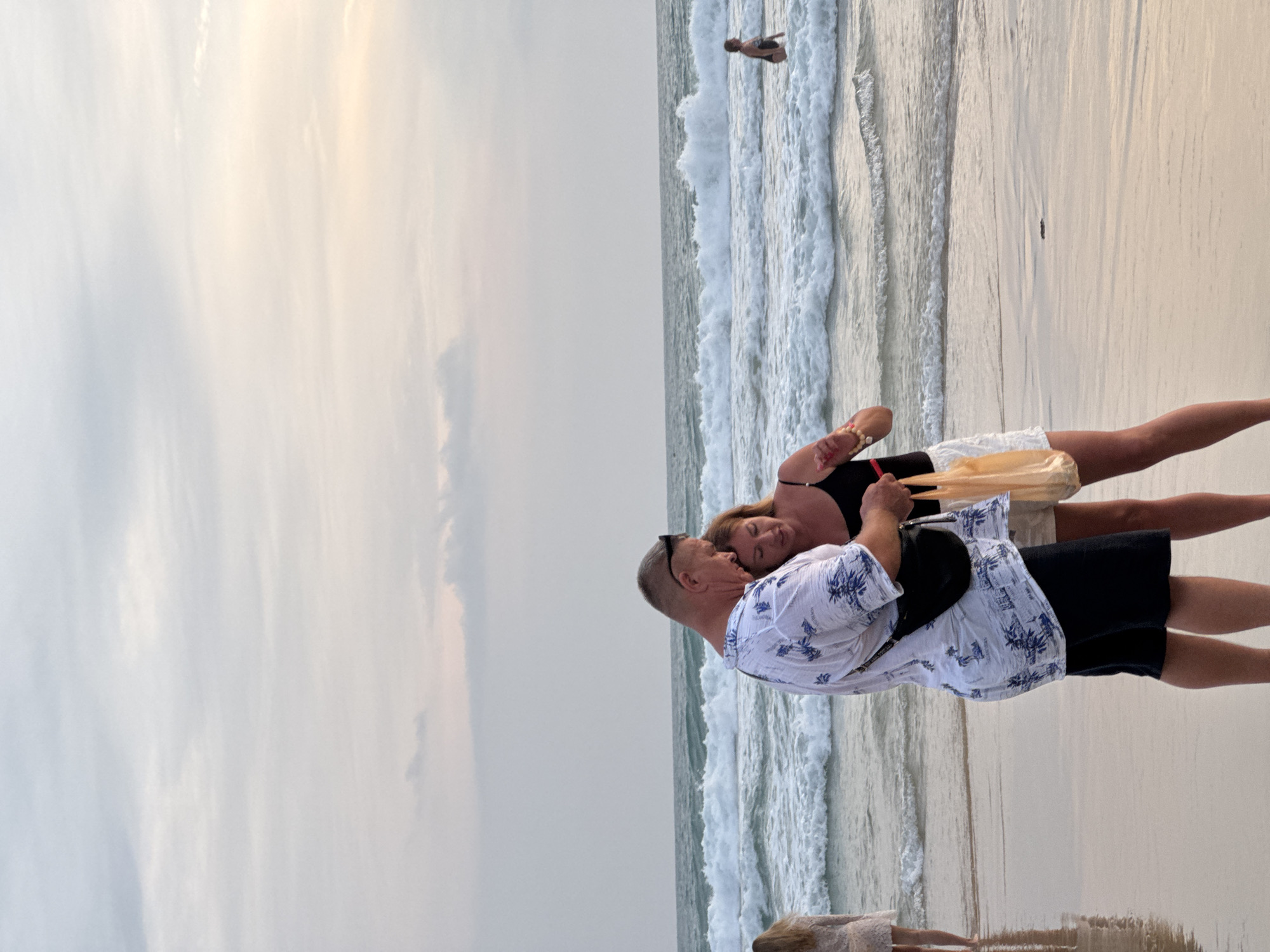 In Karon, Thailand, a man and a woman stand on a beach, facing each other. The man, wearing a white patterned shirt and dark shorts, has a black fanny pack around his waist. The woman, dressed in a black tank top and white shorts, holds a yellow bag. They are looking at something in the woman's hand. The ocean with waves stretches behind them, and other people are visible in the water and further down the beach. The sky above is cloudy.