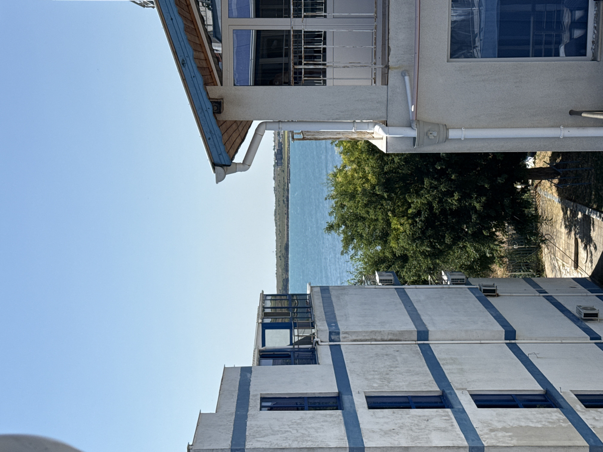 The image shows a view of the sea from an elevated position in Pomorie, Bulgaria. Two buildings, likely apartment complexes, flank the foreground. The building on the left is white with horizontal blue stripes and blue-framed windows. It has multiple floors, with a glass-enclosed balcony on the upper level. The building on the right has a white facade with a blue-trimmed roof and a balcony with a white railing. A large green tree is visible between the buildings, partially obscuring a path and a fence behind it. The sea stretches across the middle ground, with a distant, low-lying coastline. The sky is clear and blue. The overall impression is one of a sunny day in a coastal town.