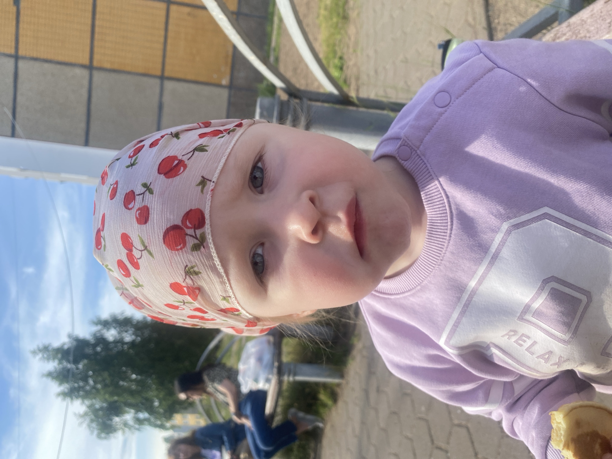 A young child is pictured in close-up, facing forward, wearing a light purple sweatshirt with "RELAX" written in white on the front and a sheer, pink headscarf with a cherry pattern. The child's eyes are looking directly at the camera. In the background, two other individuals are seated on a park bench. One person on the left is wearing a blue jacket and jeans, and the other on the right is wearing a patterned top and dark pants. A white plastic bag rests on the bench between them. The setting appears to be an outdoor park or public space, with paved ground, grass, trees, and a building with beige and yellow tiled siding visible in the distance. The sky is blue with some clouds. The scene is likely in the city of Chaikovsky, Russia, given the context.
