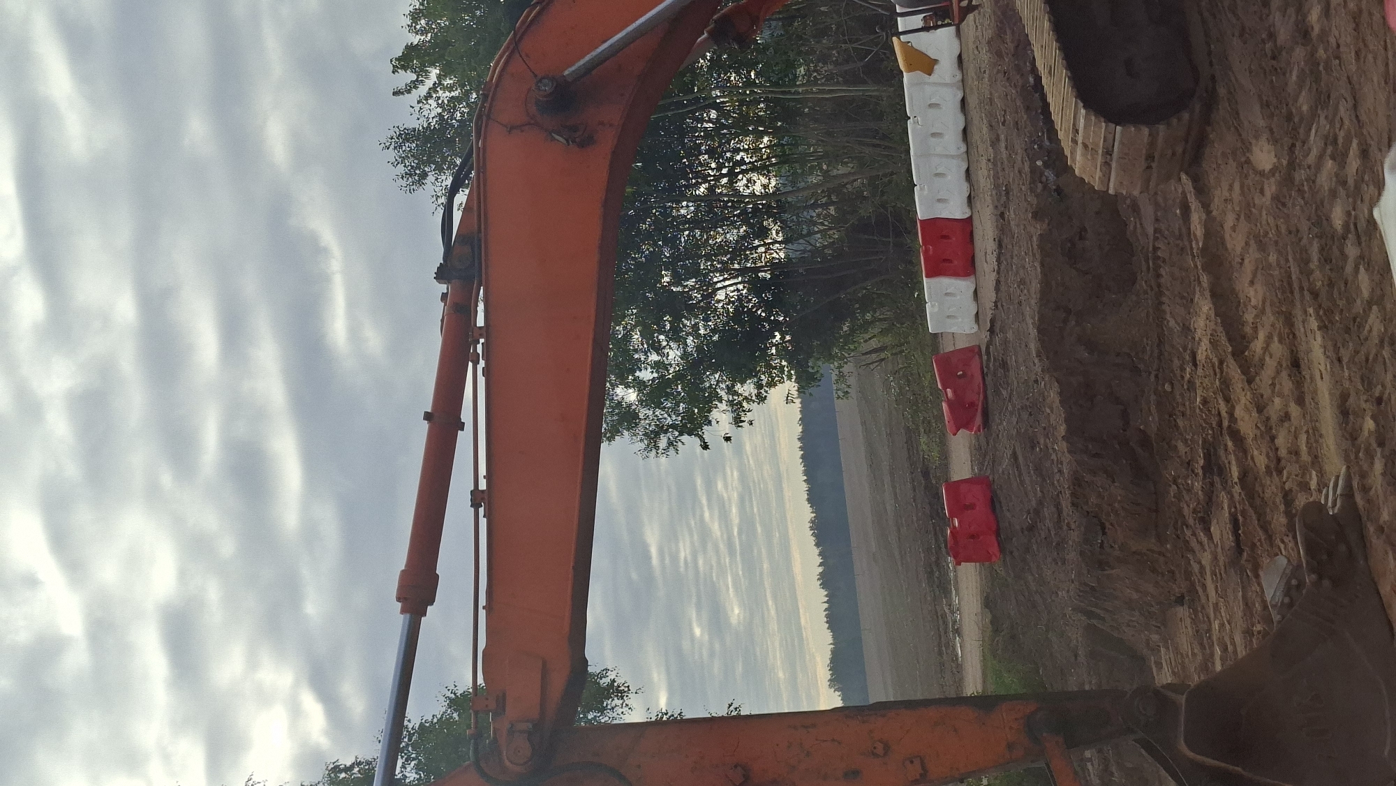 An orange excavator arm, with a visible hydraulic cylinder and bucket, is positioned in the foreground of a construction site. The ground is composed of disturbed soil, with tire tracks visible. In the middle ground, a line of red and white plastic barrier blocks demarcates an area. Behind the barriers, a line of trees forms the background, with a field and a distant treeline under a cloudy sky. The scene is set in the vicinity of Бягомльскі сельскі Савет, Belarus.