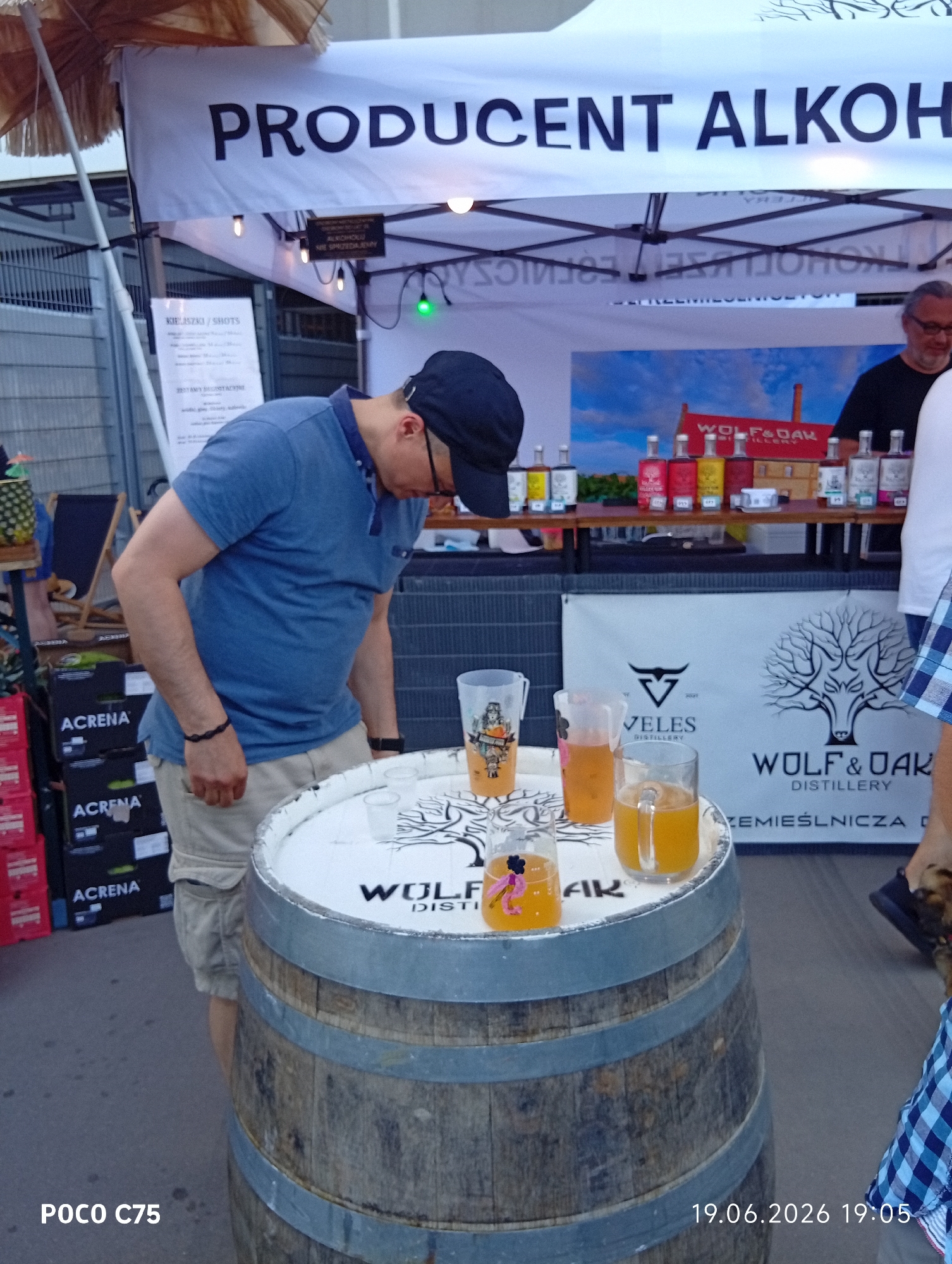 A person in a blue polo shirt and beige shorts is standing by a wooden barrel on which four glasses of amber-colored liquid and a small decorative item are placed. The person is looking down. Behind the barrel is a white banner with text in Polish, including "PRODUCENT ALKOHOL..." and logos for "VELES DISTILLERY" and "WOLF & OAK DISTILLERY". On a shelf behind the banner are several bottles of alcohol. In the background, a man in a black shirt and glasses is visible. The setting appears to be an outdoor market or event. The timestamp "19.06.2026 19:05" is visible in the bottom right corner, and "POCO C75" is in the bottom left. The location is identified as Wrocław, Poland.
