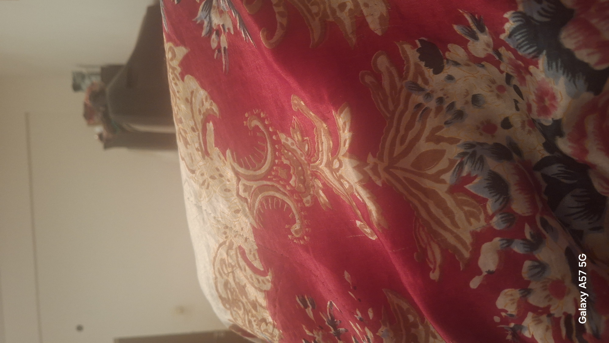 The image displays a close-up of a red fabric with a floral and paisley-like pattern. The pattern features intricate designs in gold and cream, with accents of blue and pink flowers. The fabric appears to be a blanket or bedspread, draped over an unseen surface, creating folds and highlights that suggest a soft texture. In the upper background, a blurred out-of-focus area shows a wall and a hint of furniture, possibly in a room. The bottom left corner of the image contains the text "Galaxy A57 5G". There are no people or specific city details visible in the image.